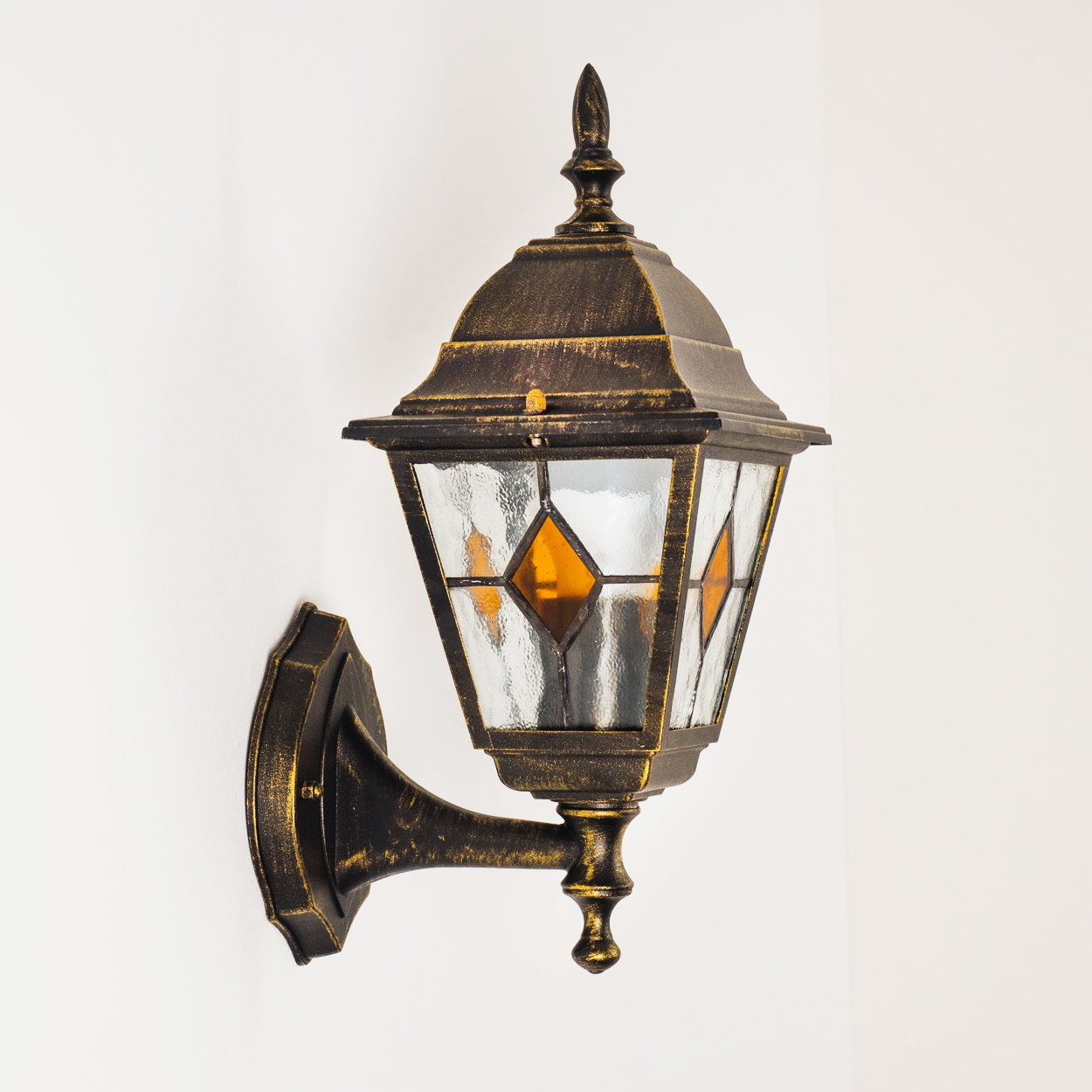 Eine bronzefarbene, an der Wand befestigte Laterne mit Glasscheiben und einer bernsteinfarbenen Raute. Der Hintergrund ist eine schlichte weiße Wand.