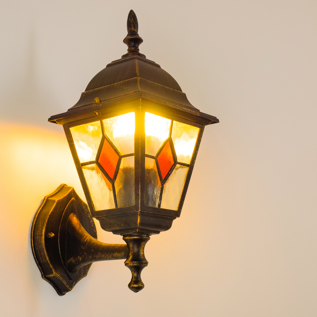 Eine verzierte Außenlampe mit Buntglasscheiben und einem goldfarbenen Rahmen. Sie ist an einer weißen Wand mit warmem Glanz befestigt.