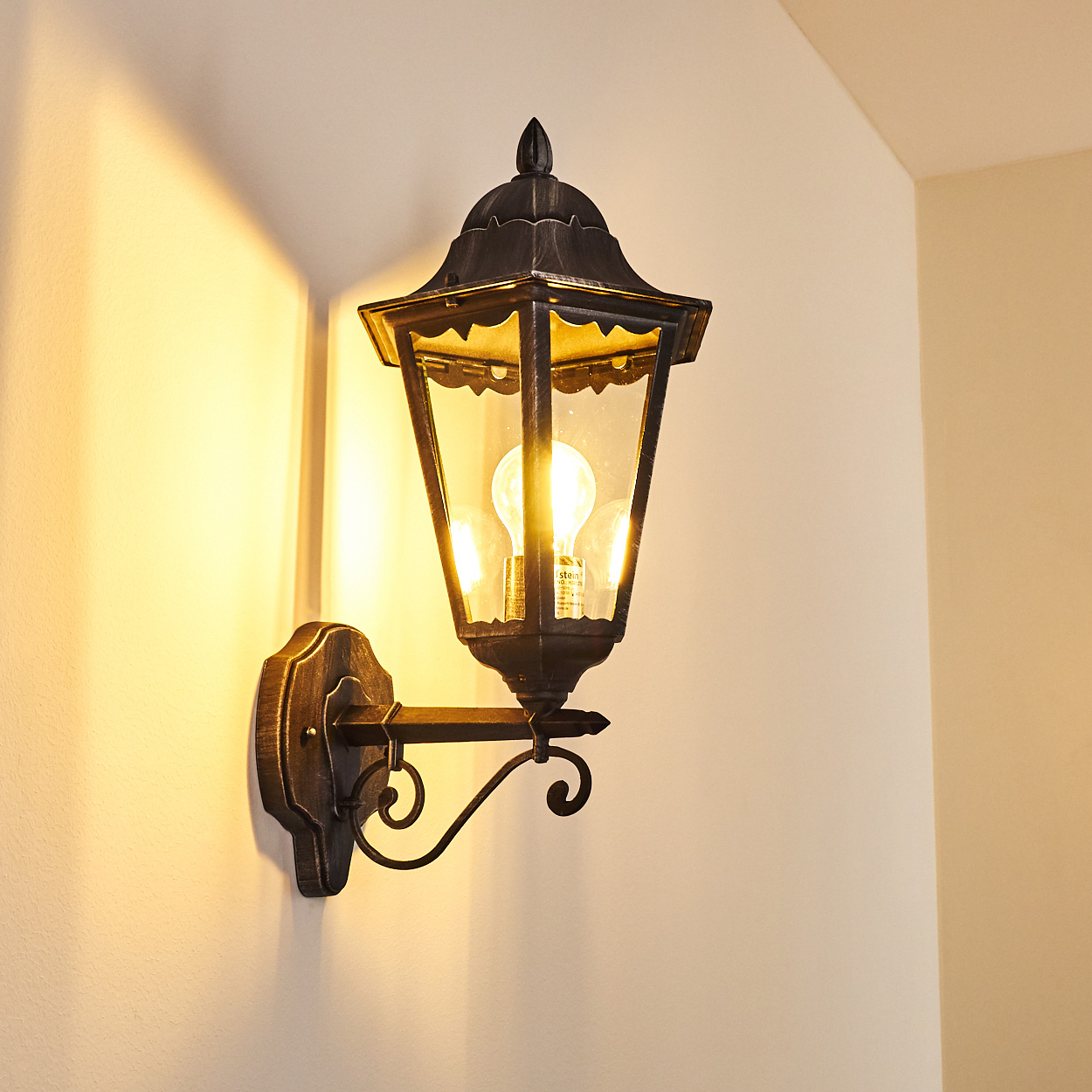 Eine dekorative Außenwandleuchte mit dunklem Metall-Finish. Die Lampe leuchtet und wirft einen warmen Schein auf die weiße Wand.