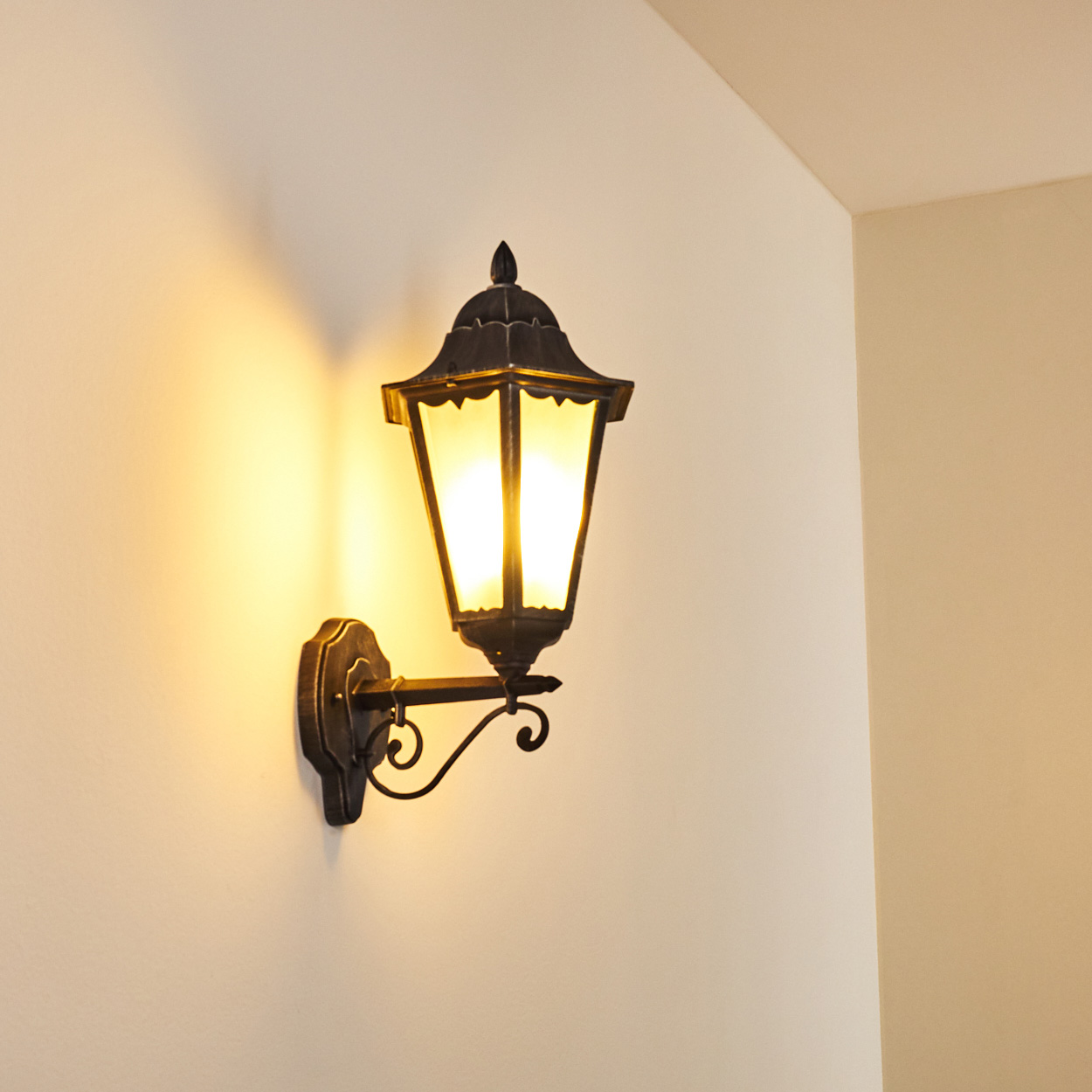 Eine verzierte, schwarz an der Wand montierte Lampe mit leuchtendem, gelb getöntem Leuchtmittel an cremefarbener Wand. Ecke einer weiteren Wand sichtbar.