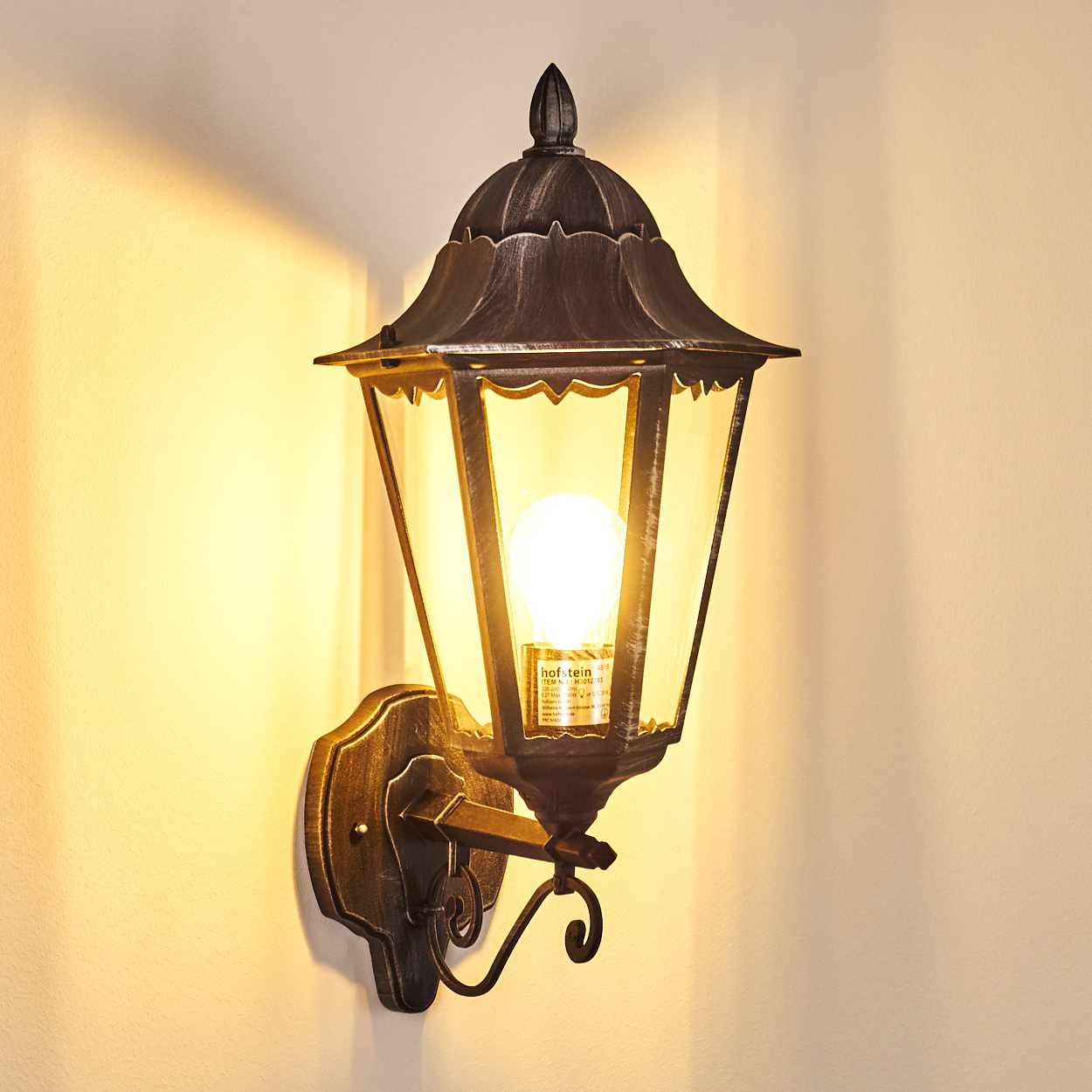 Eine Außenwandlampe mit einer leuchtenden Glühbirne. Sie hat einen dunklen Metallrahmen und klare Glasscheiben. Die Wand dahinter ist hellbeige.