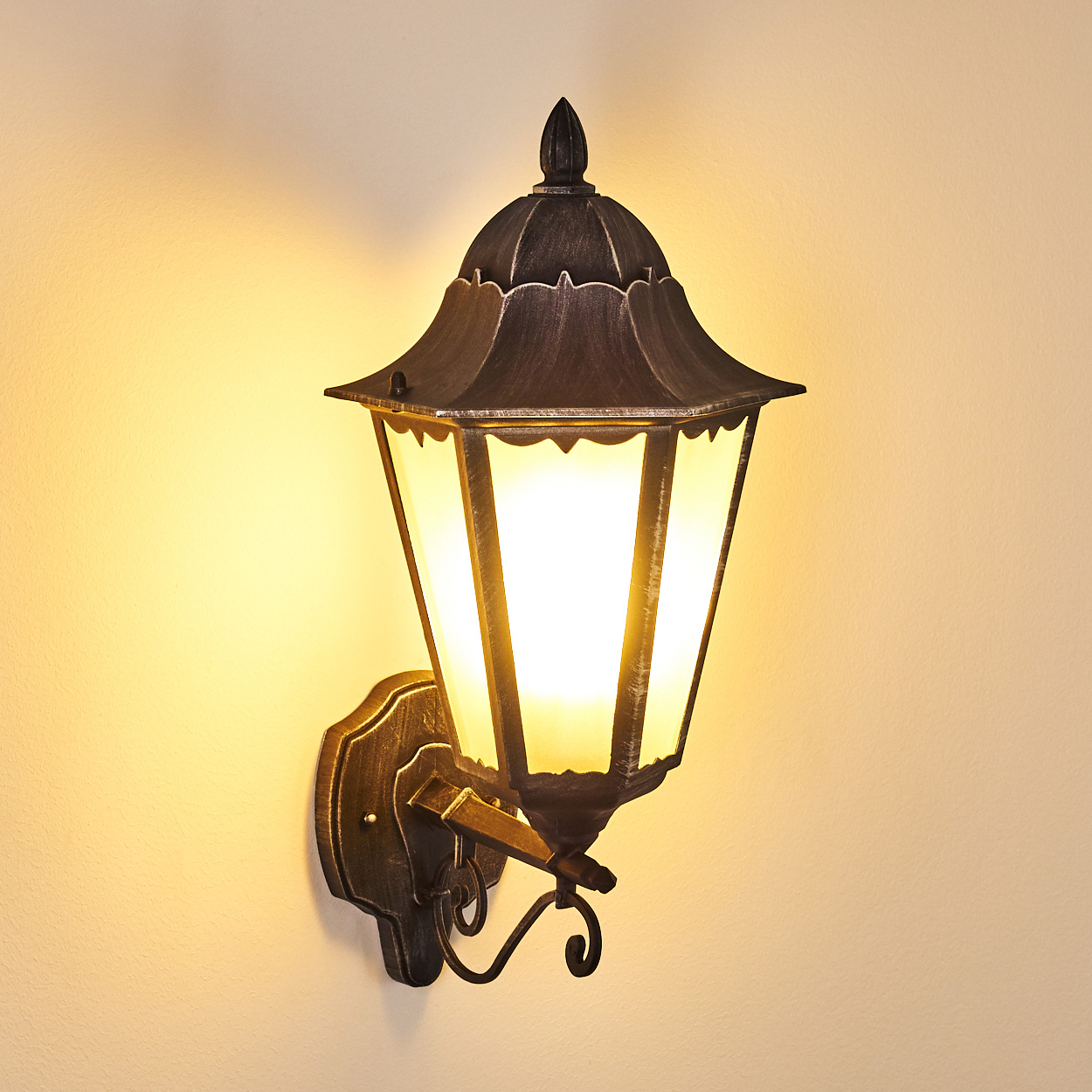 Eine verzierte, an der Wand befestigte Laterne mit dunklem Metall-Finish. Sie hat ein warmes Licht und steht vor einer cremefarbenen Wand.