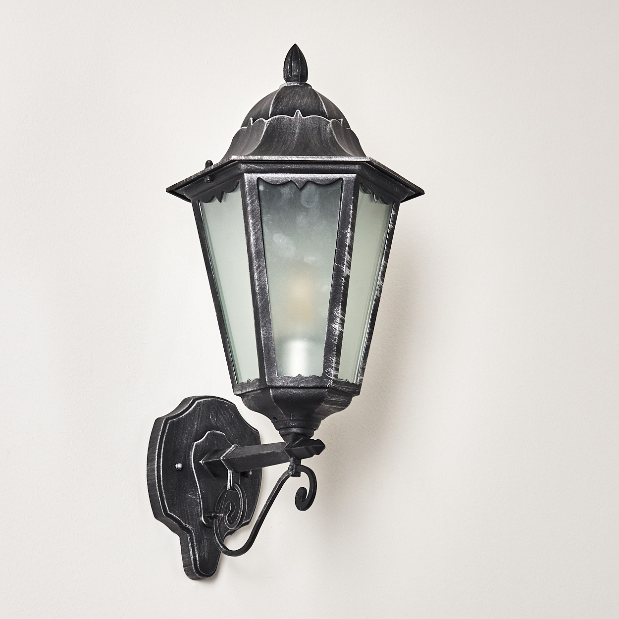 Eine schwarze und silberne Außenlampe ist an einer weißen Wand befestigt. Die Lampe hat ein Glasgehäuse und eine dekorative Halterung.