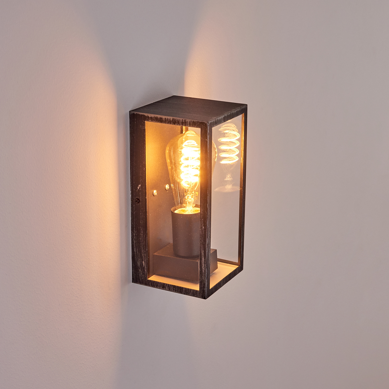 Eine rechteckige, schwarze Wandleuchte mit Glasgehäuse. Im Inneren ist eine leuchtende Glühbirne sichtbar. Die Lampe ist an einer weißen Wand montiert.
