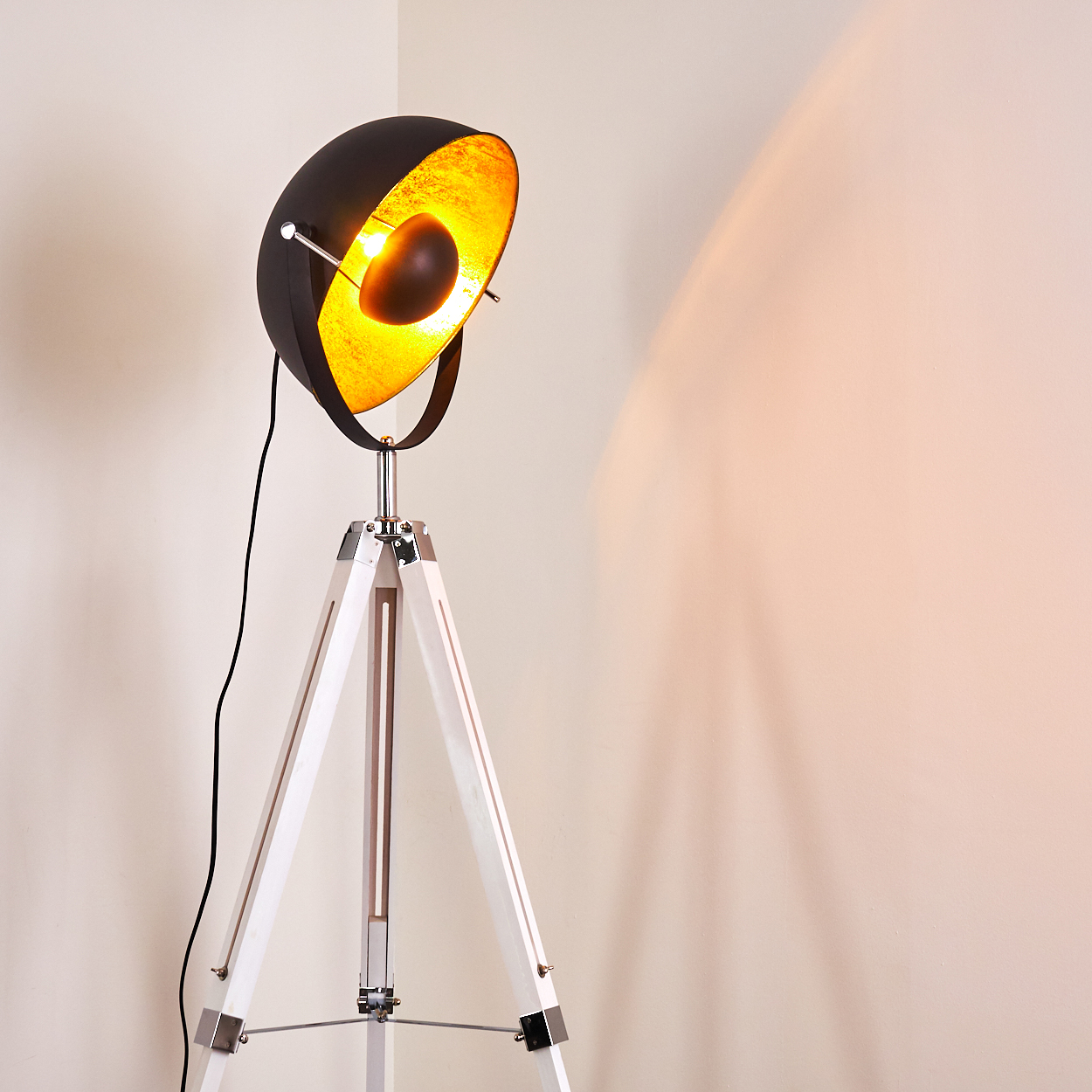 Eine schwarz-goldene Stehlampe mit weißem Stativfuß. Die Lampe wirft einen warmen Schein an die Wand. Das Kabel ist schwarz.