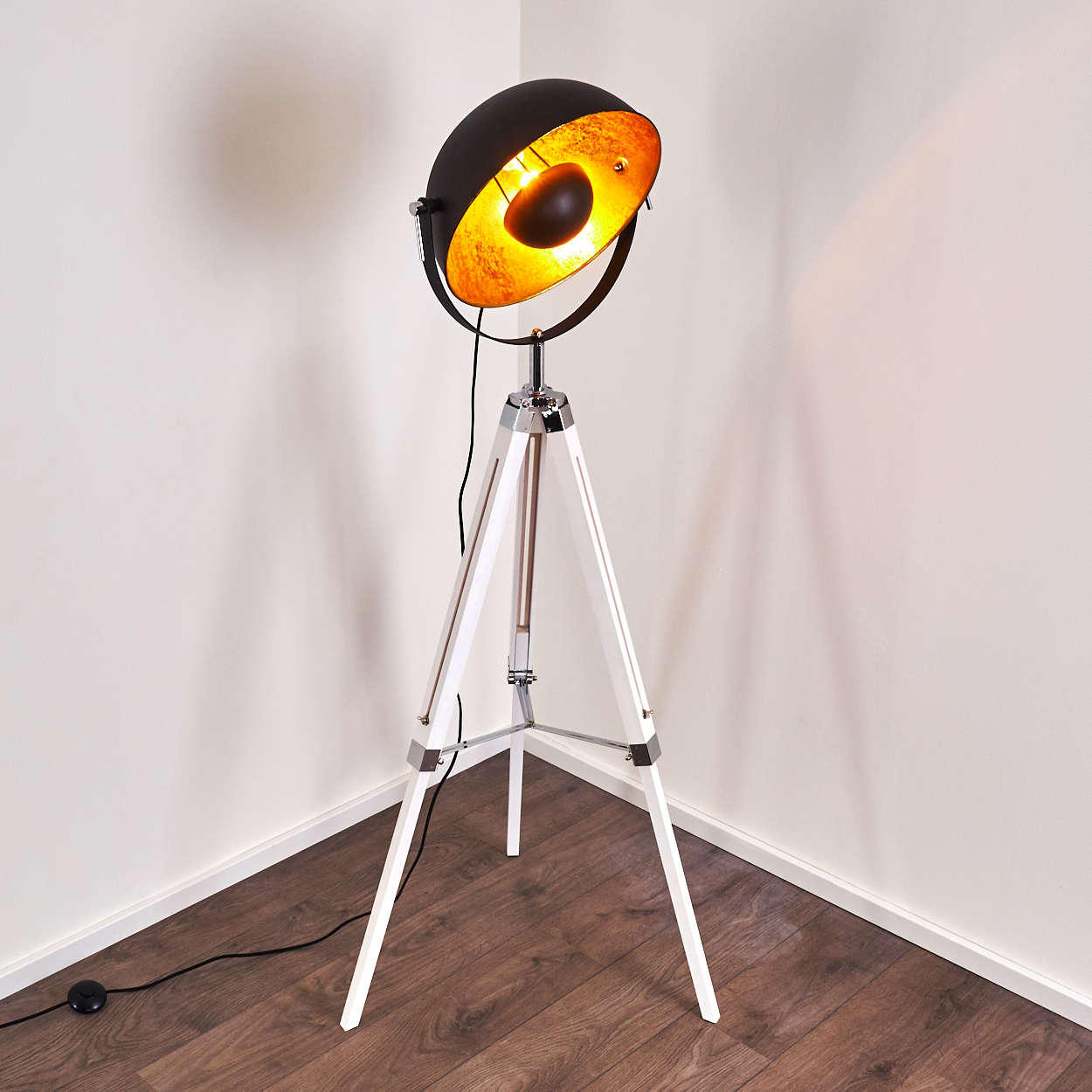 Eine schwarz-goldene Stehlampe mit weißem Dreibein-Fuß steht in einer Ecke. Im Hintergrund sind Holzboden und weiße Wände zu sehen.