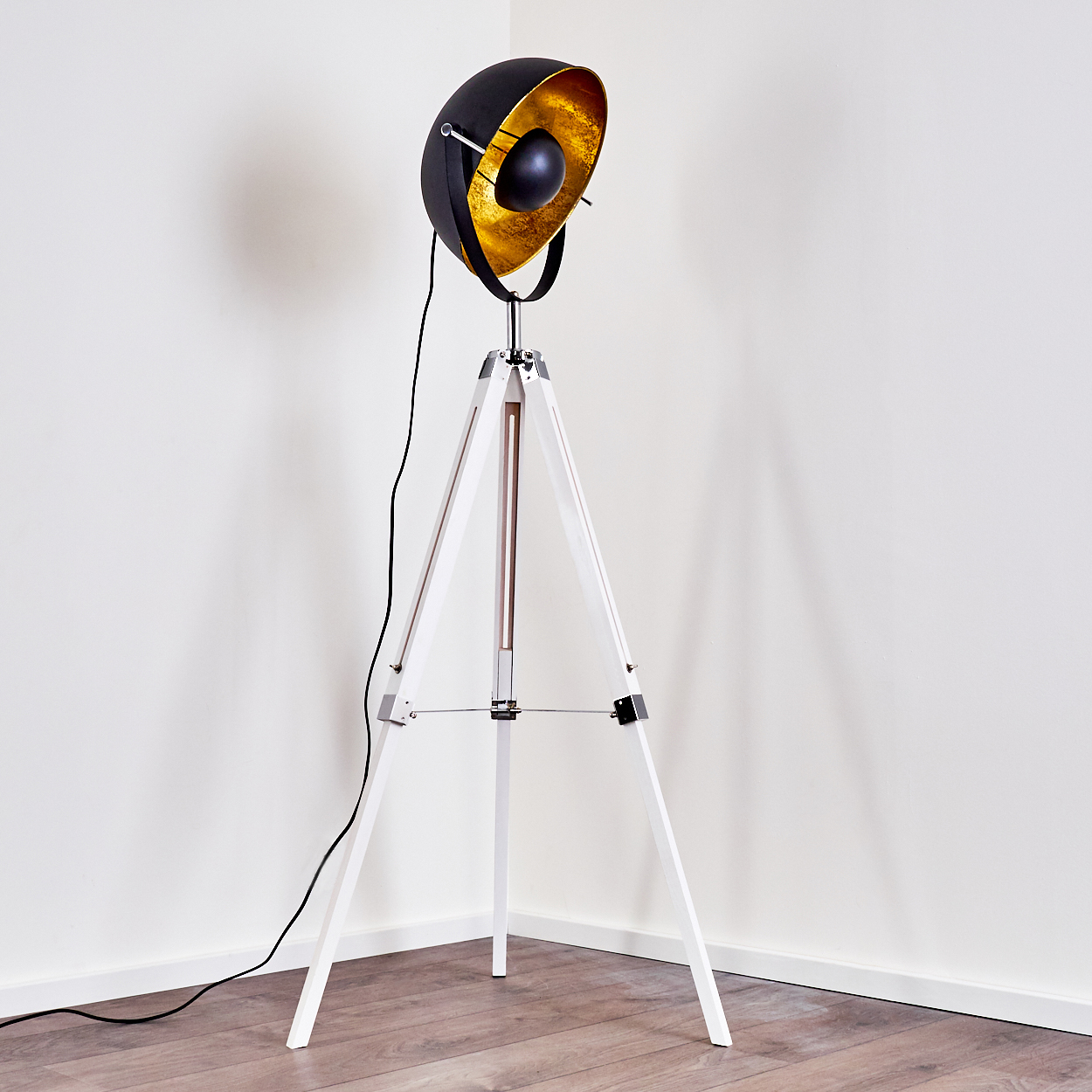 Eine schwarze und goldene Scheinwerferlampe auf einem weißen Stativ, das in einer Ecke auf einem Holzboden vor einer weißen Wand steht.