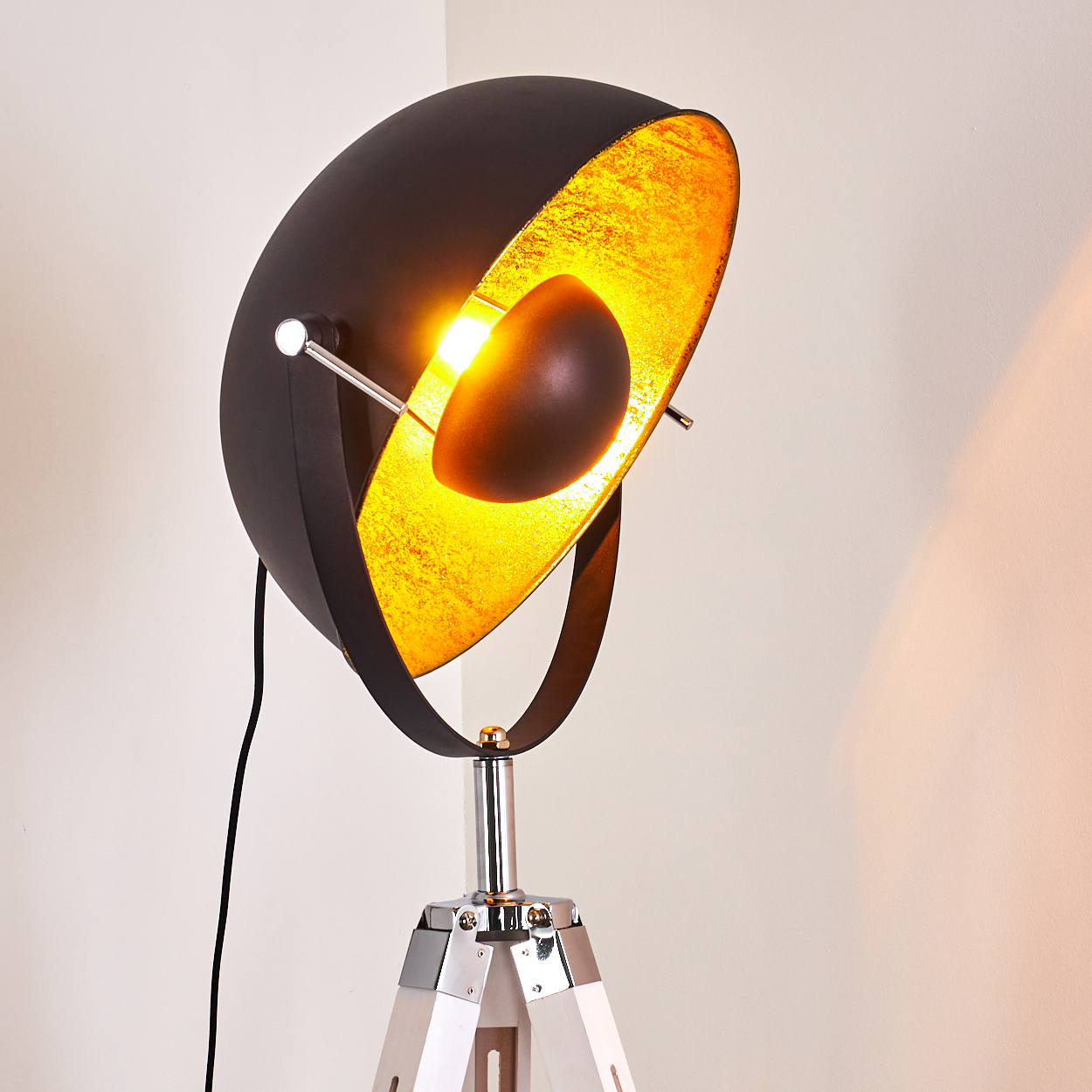 Eine schwarz-goldene Scheinwerferlampe mit Stativfuß. Das Licht ist an und beleuchtet das goldene Innere. Weiße Wand im Hintergrund.