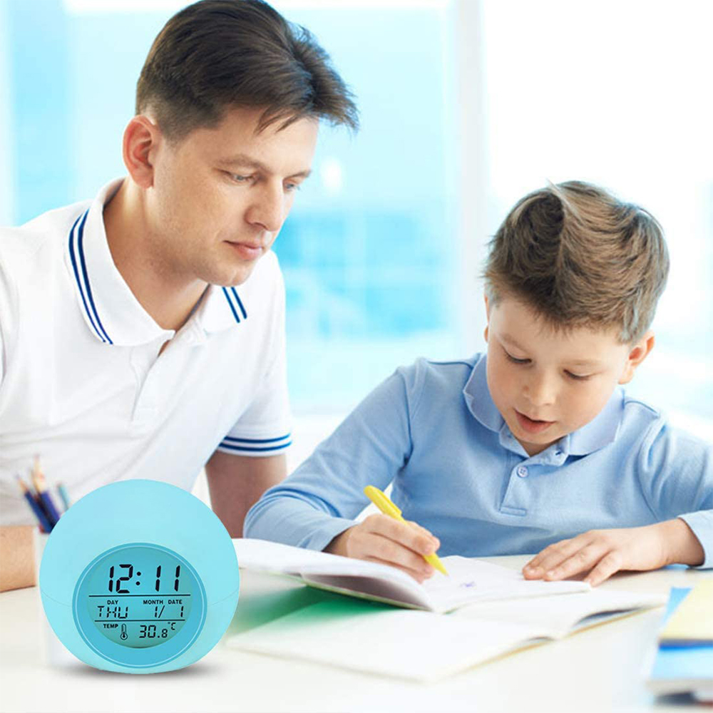 Ein Vater beobachtet seinen Sohn bei den Hausaufgaben. Eine blaue Uhr zeigt 12:11. Der Junge schreibt mit einem gelben Stift.