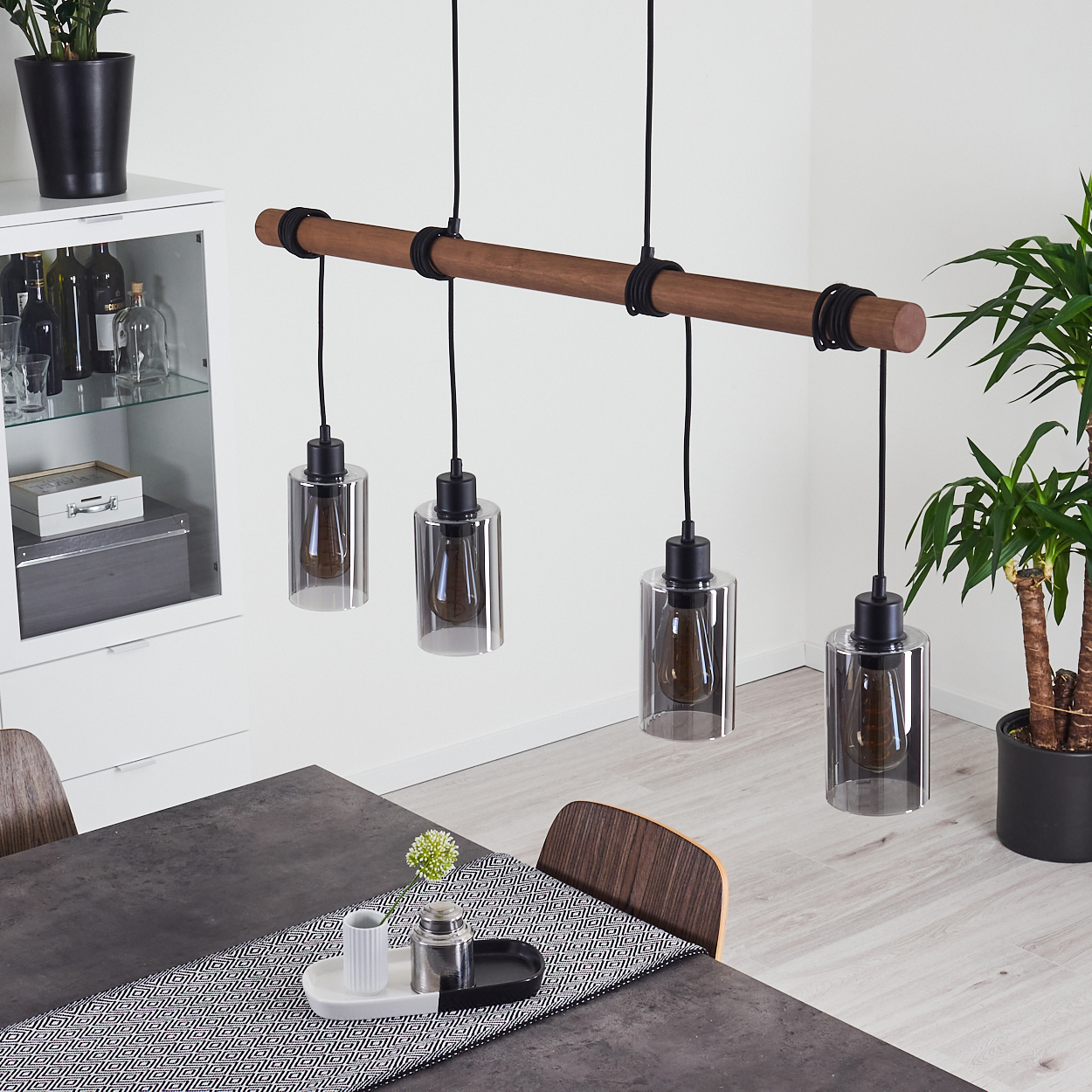 Ein modernes Esszimmer mit einer Deckenleuchte aus Holzbalken. Ein Tisch ist mit einem Läufer und einem Mittelstück gedeckt. Eine Pflanze steht in der Ecke.