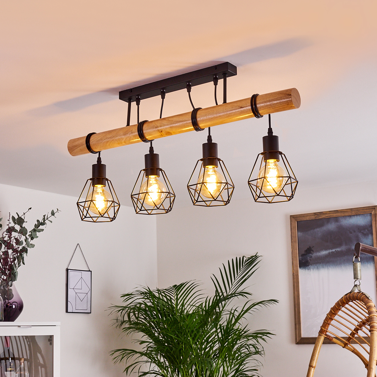 Ein Holzbalken mit vier Lampen im Käfigstil hängt von der Decke. Im Hintergrund befinden sich eine grüne Pflanze und ein gerahmtes Bild.
