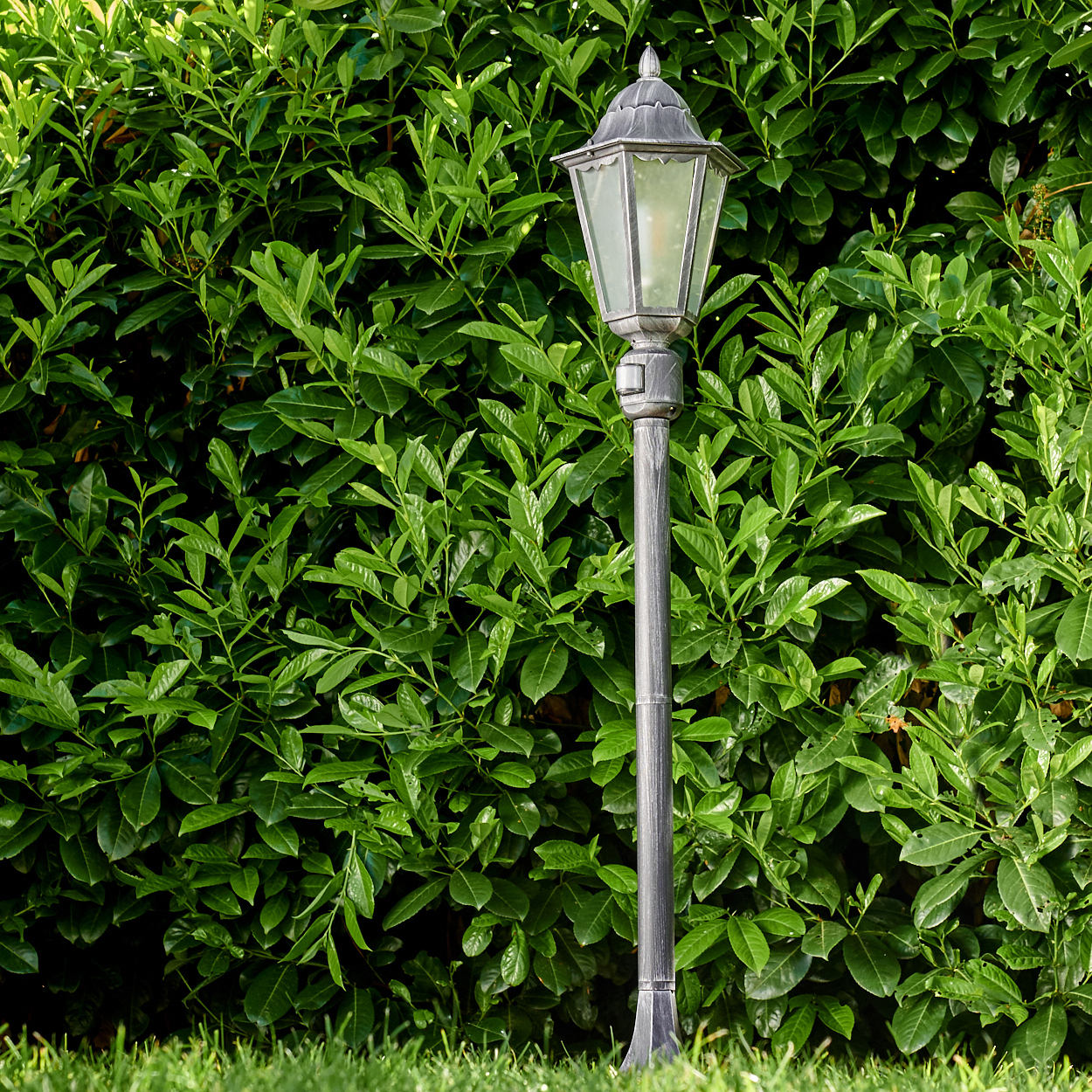 Eine graue Straßenlaterne steht vor einer dichten, grünen Hecke. Im Vordergrund ist grünes Gras zu sehen. Die Lampe ist verziert, mit Glasscheiben.
