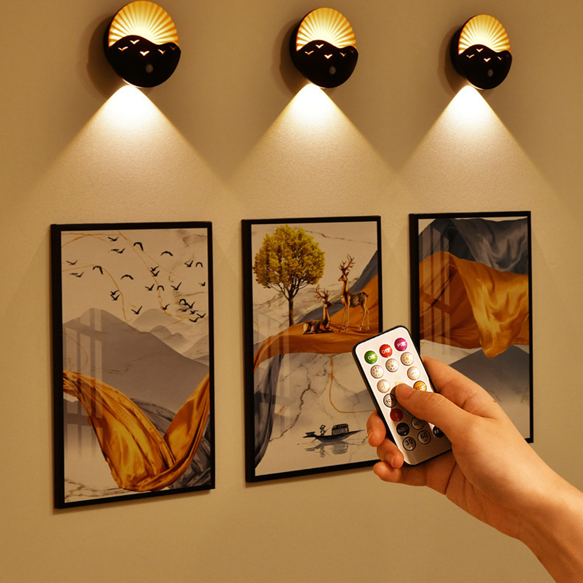 Wandkunst mit drei beleuchteten Lampen über gerahmten Gemälden. Eine Hand hält eine Fernbedienung. Gemälde zeigen Landschaften und Rehe.