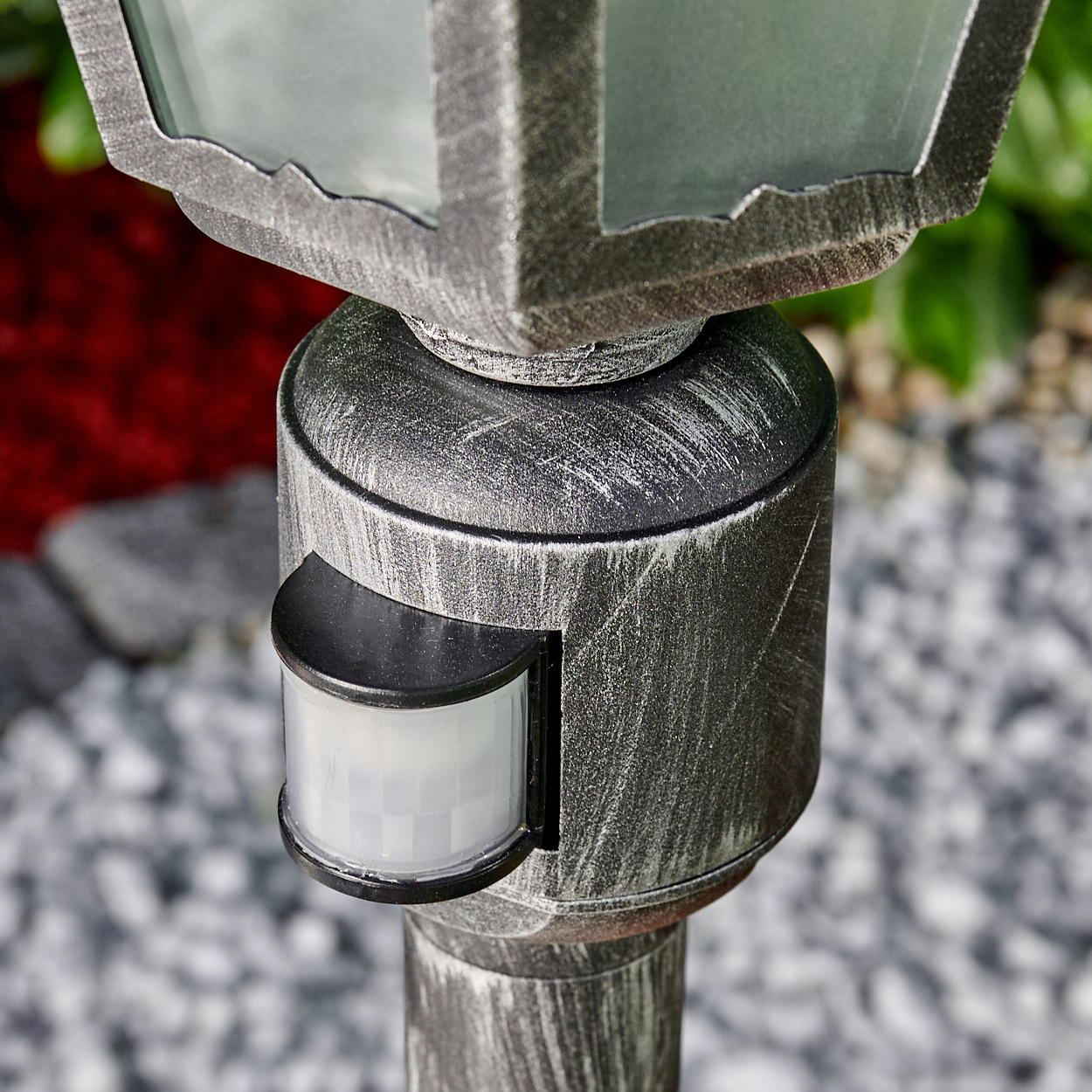 Nahaufnahme einer metallischen Außenlampe mit Bewegungssensor. Die Lampe hat einen quadratischen Kopf und steht auf einem Pfosten. Roter und grüner Hintergrund.