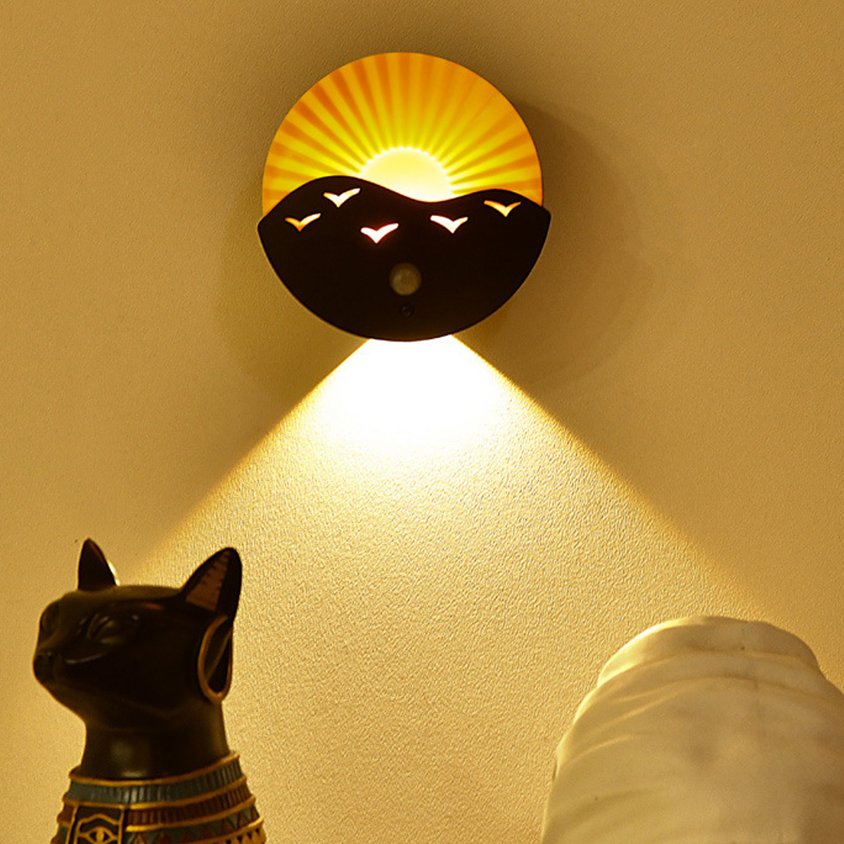 Eine Wandlampe mit Sonnendesign beleuchtet die Wand. Eine schwarze Katzenstatue und ein weißes Objekt befinden sich darunter.