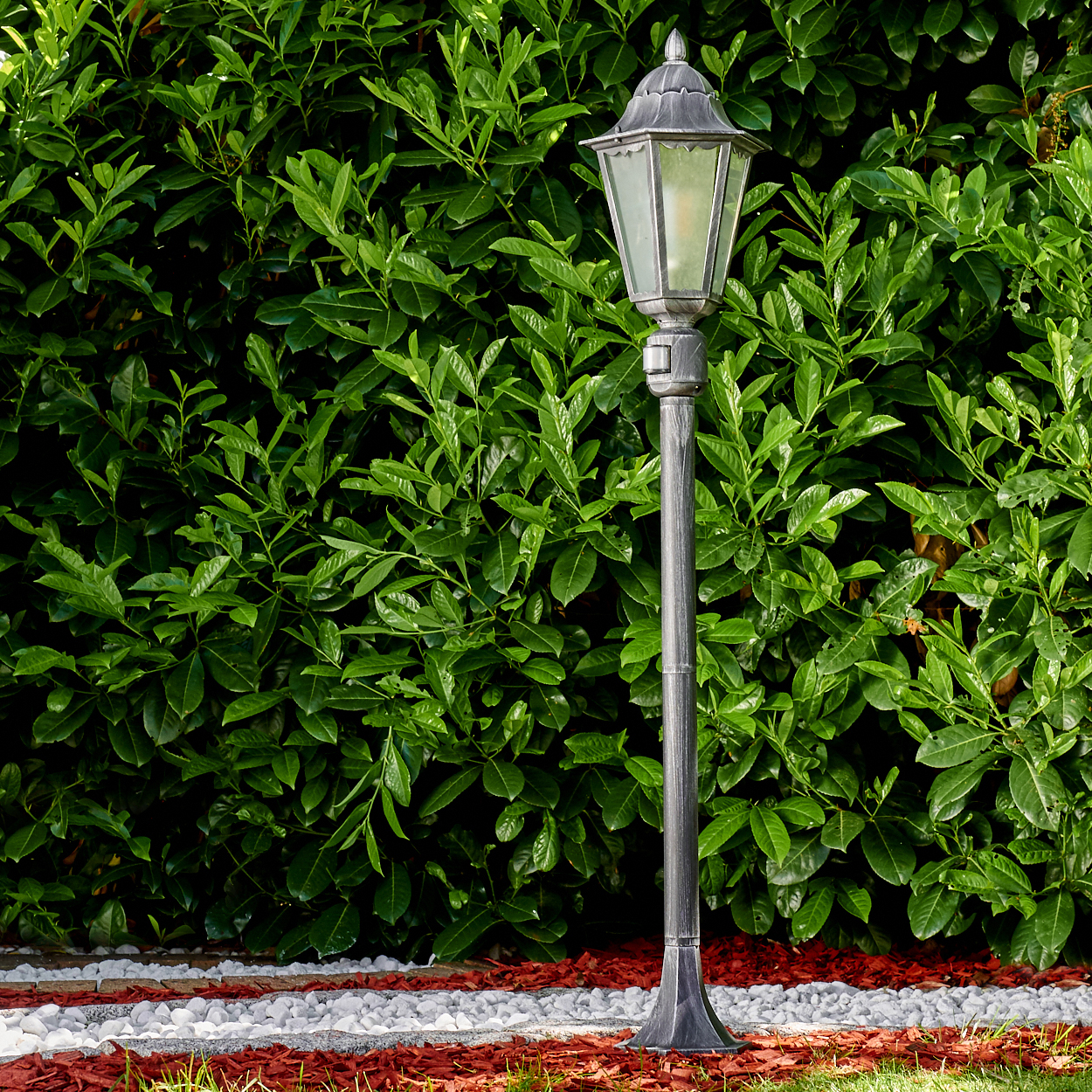 Eine metallische Straßenlaterne steht vor einer dichten, grünen Hecke. Der Boden zeigt roten und weißen Kies und grünes Gras.