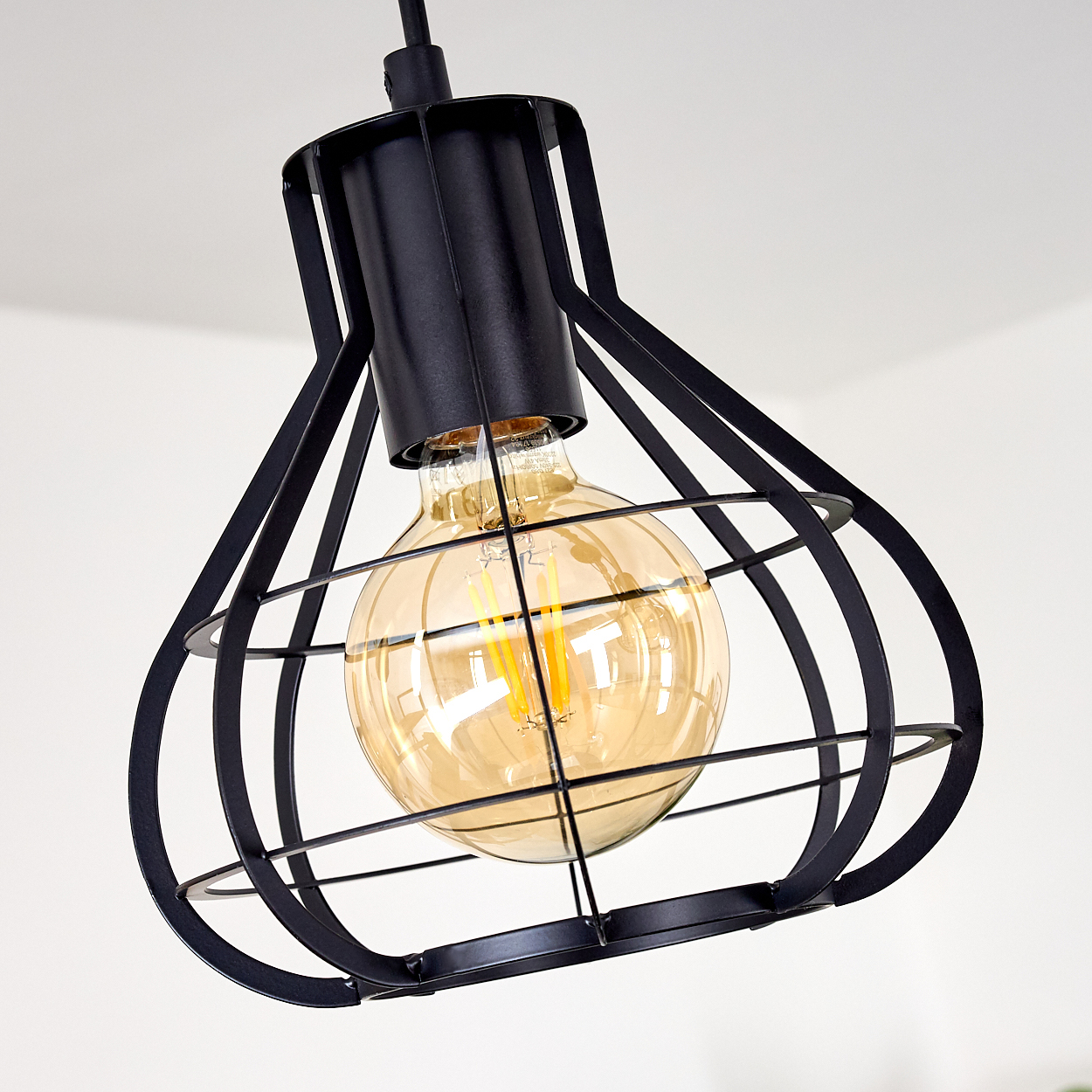 Eine schwarze Metallkäfig-Leuchte mit einer runden, bernsteinfarbenen Glühbirne vor einer weißen Decke.