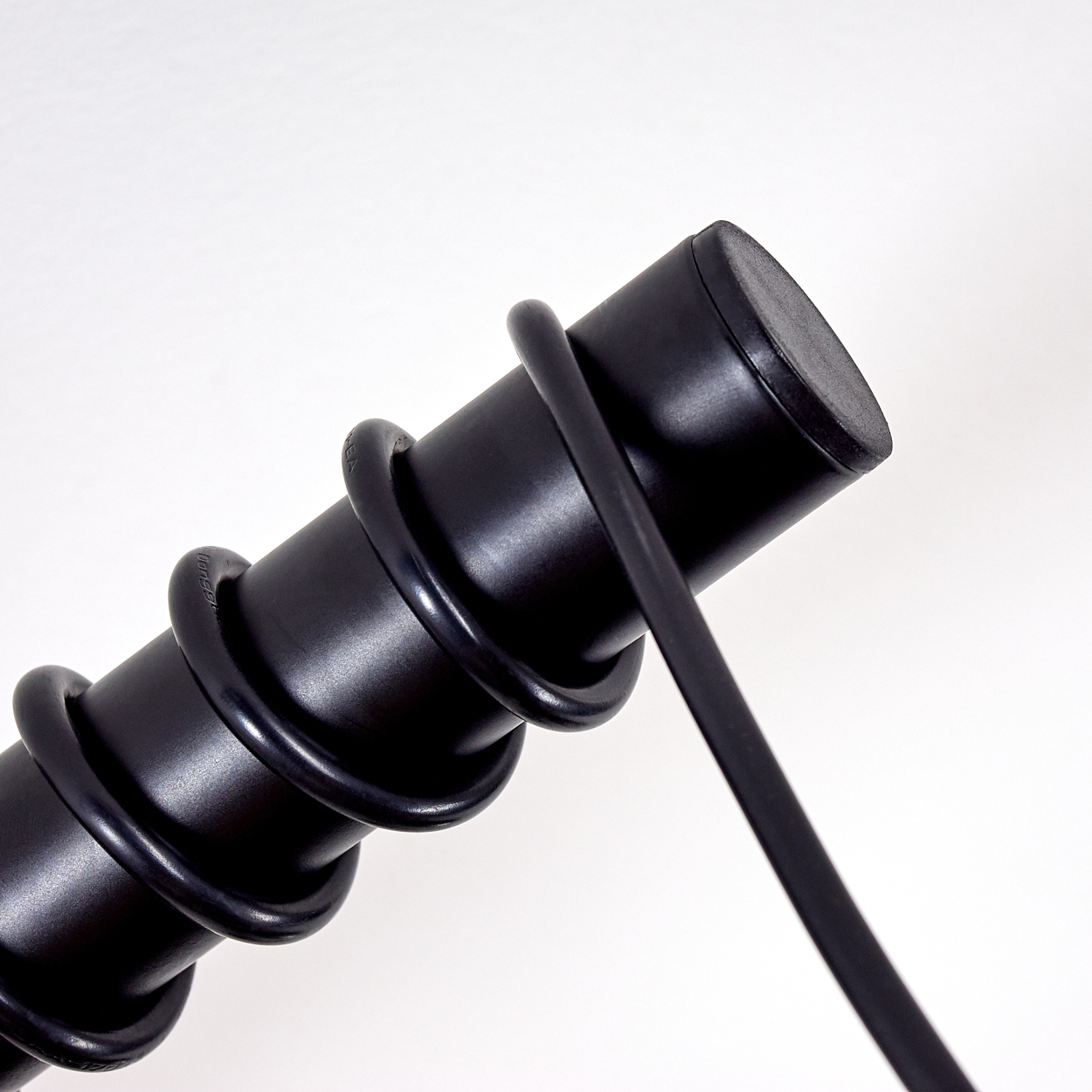 Nahaufnahme eines schwarzen zylindrischen Objekts, das mit einer schwarzen Spirale umwickelt ist. Ein schwarzer flacher Riemen erstreckt sich von der Seite. Weißer Hintergrund.