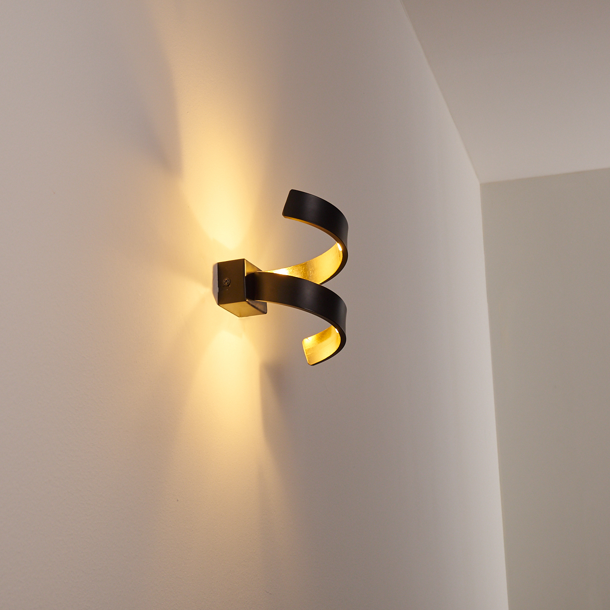 Eine Wandlampe mit schwarzem, spiralförmigem Design und goldenem Innenraum, montiert an einer weißen Wand, die warmes Licht wirft.