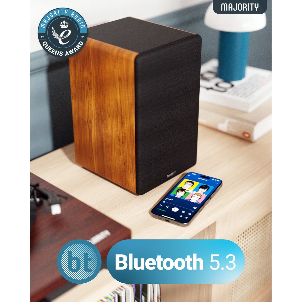 Ein Lautsprecher mit Holzseite, ein Musik abspielendes Telefon und ein Bluetooth 5.3 Logo stehen auf einem Tisch. Im Hintergrund eine Lampe und Bücher.