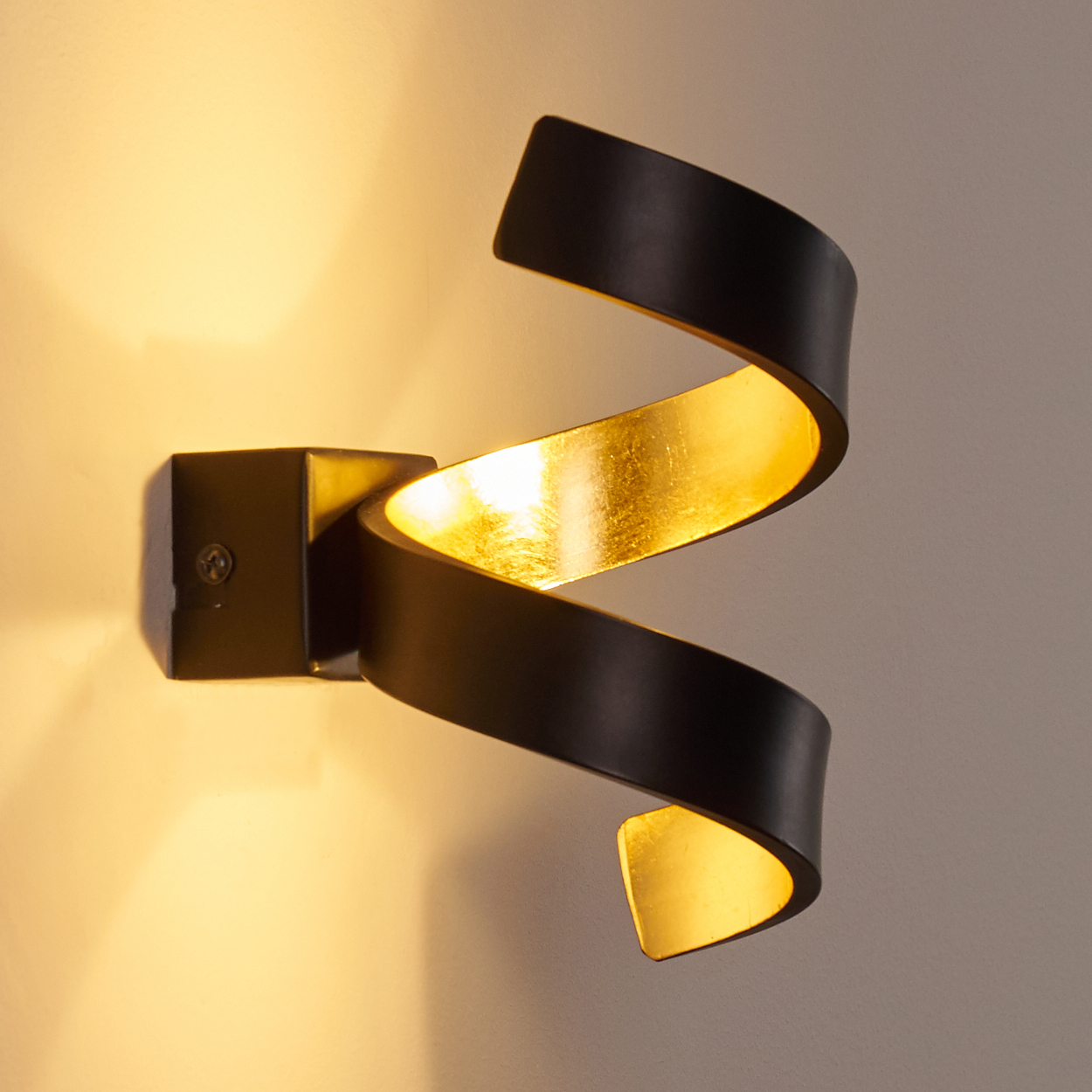 Eine schwarze, spiralförmige Wandleuchte mit goldfarbenem Innenraum ist an einer cremefarbenen Wand befestigt und wirft warmes Licht.