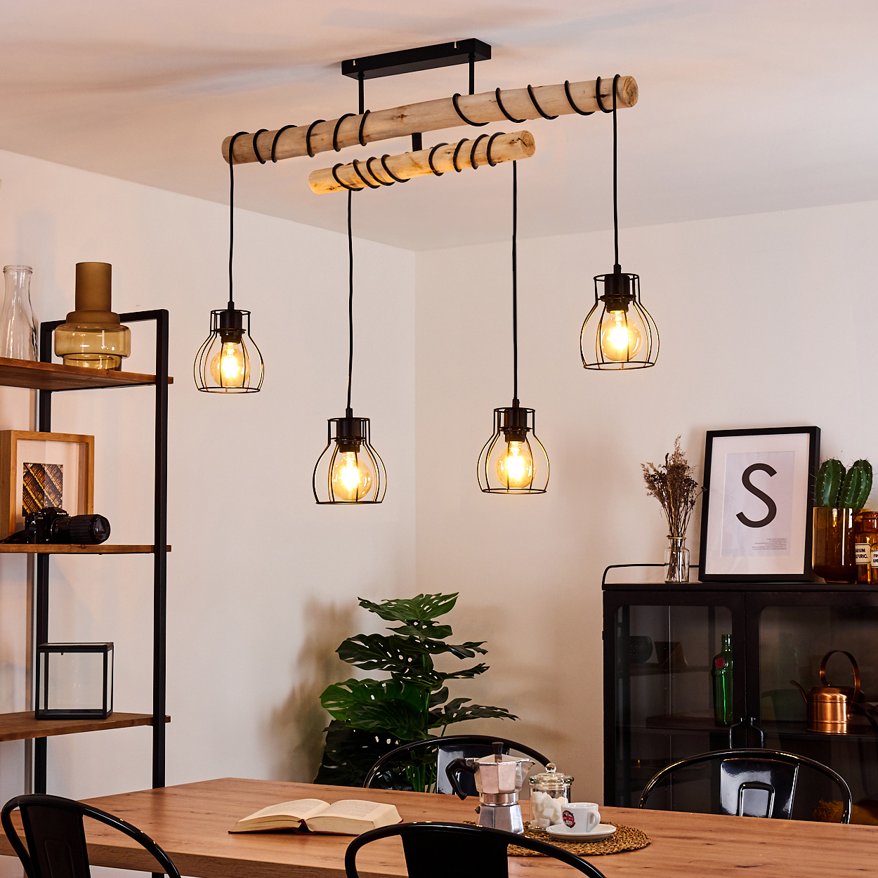 Ein Esszimmer mit Holztisch, schwarzen Stühlen und einer Hängelampe mit vier Glühbirnen. Es gibt ein Bücherregal und einen Schrank.
