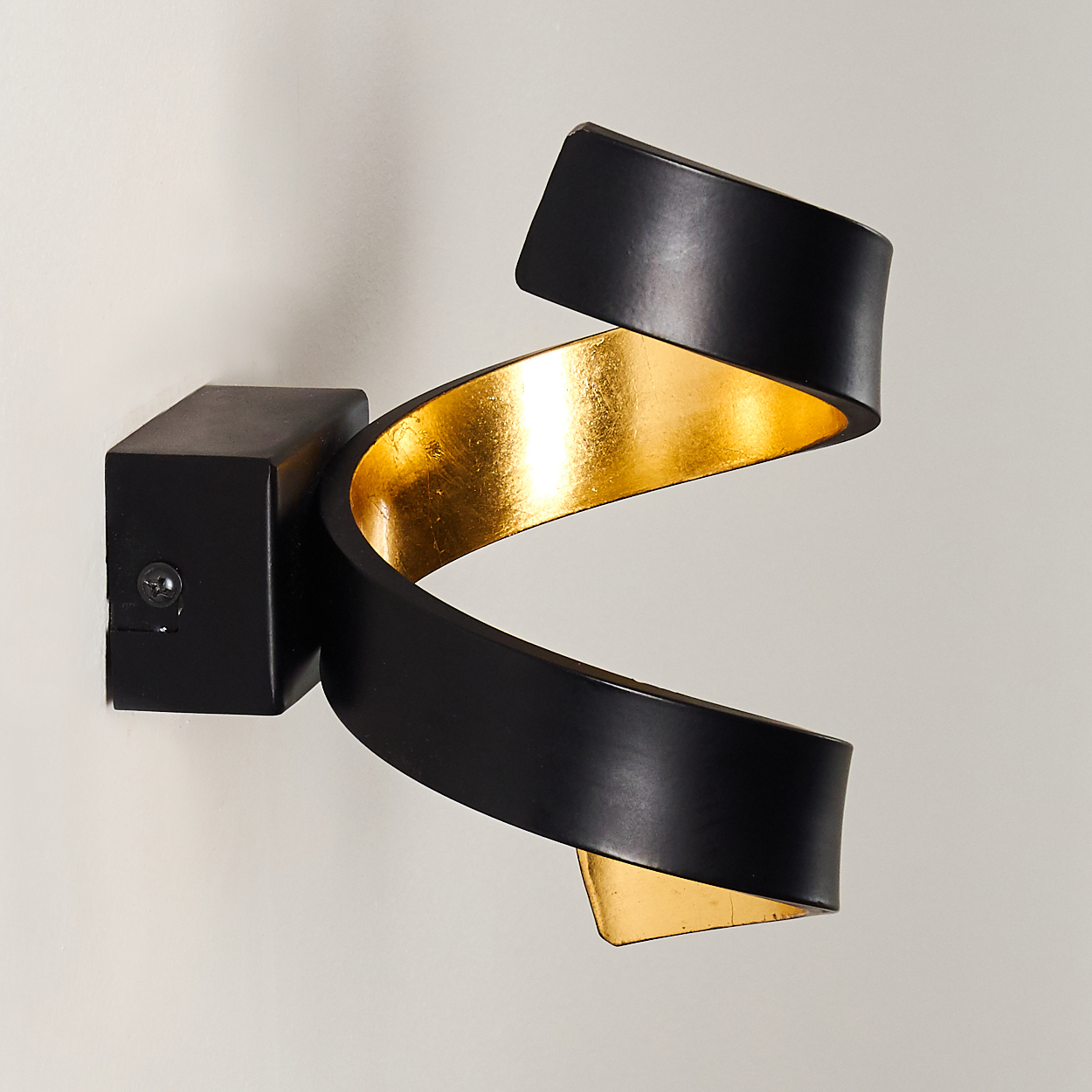Eine moderne Wandleuchte mit schwarzer Außenseite und goldener Innenseite, montiert an einer weißen Wand. Das Design ist eine wirbelnde Bandform.