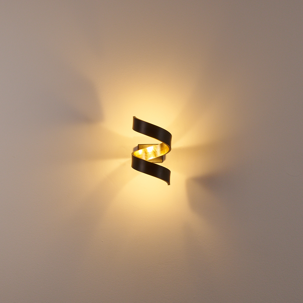Eine Wandleuchte mit Spirale. Sie wirft warmes Licht auf eine hellbeige Wand. Die Lampe hat außen Dunkelbraun und innen Gold.