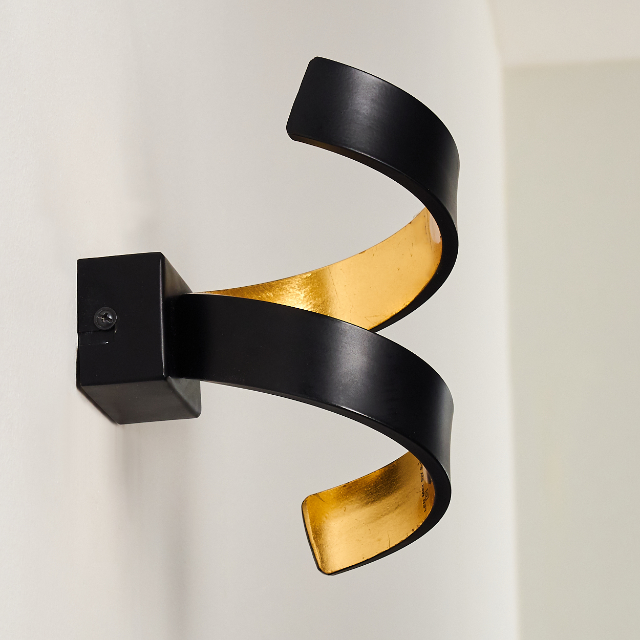 Eine schwarz-goldene Wandleuchte in Spiralform ist an einer weißen Wand befestigt. Die Leuchte hat eine quadratische Basis und ein gebogenes, bandartiges Design.