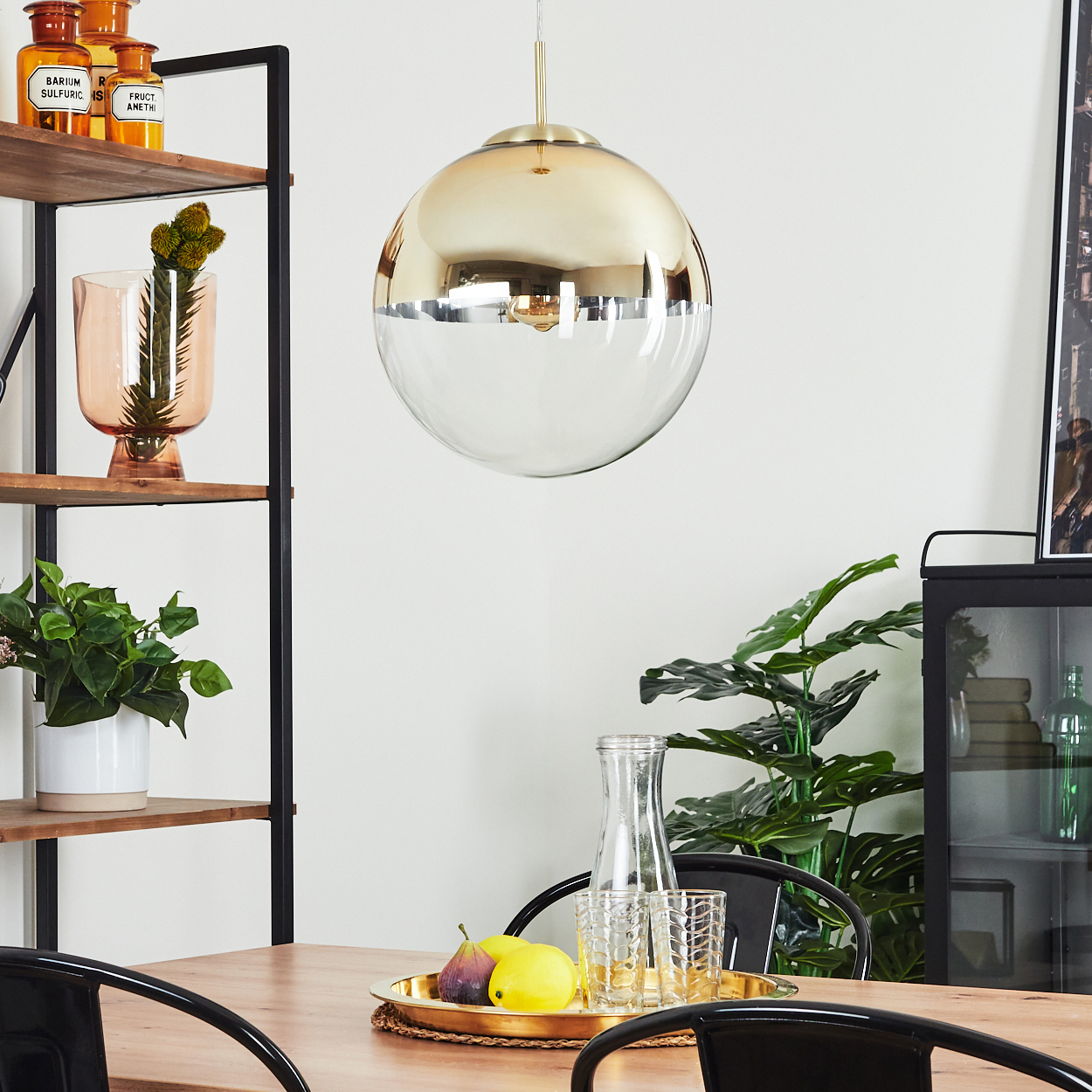 Eine goldene und gläserne Pendelleuchte hängt über einem Holztisch. Auf dem Tisch steht ein Tablett mit Obst und Gläsern. Regale und Pflanzen sind im Hintergrund.
