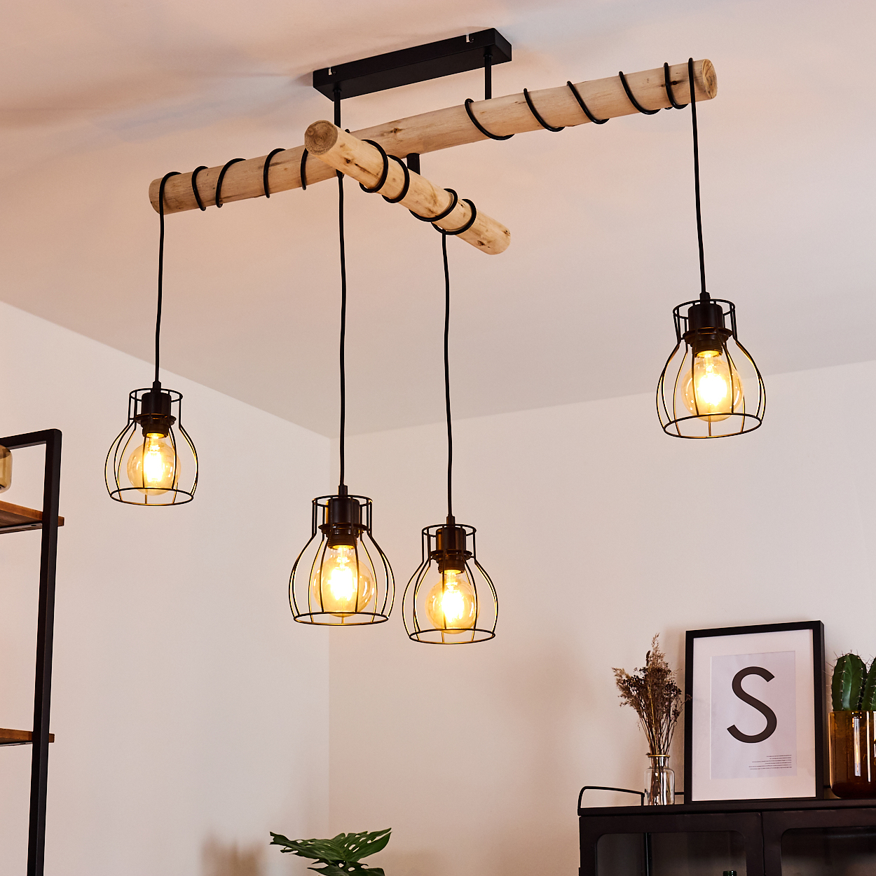 Eine moderne Deckenleuchte mit vier käfigartigen Lampen, die von Holzbalken hängen. Auf einem schwarzen Schrank befinden sich ein gerahmtes Kunstwerk und Pflanzen.