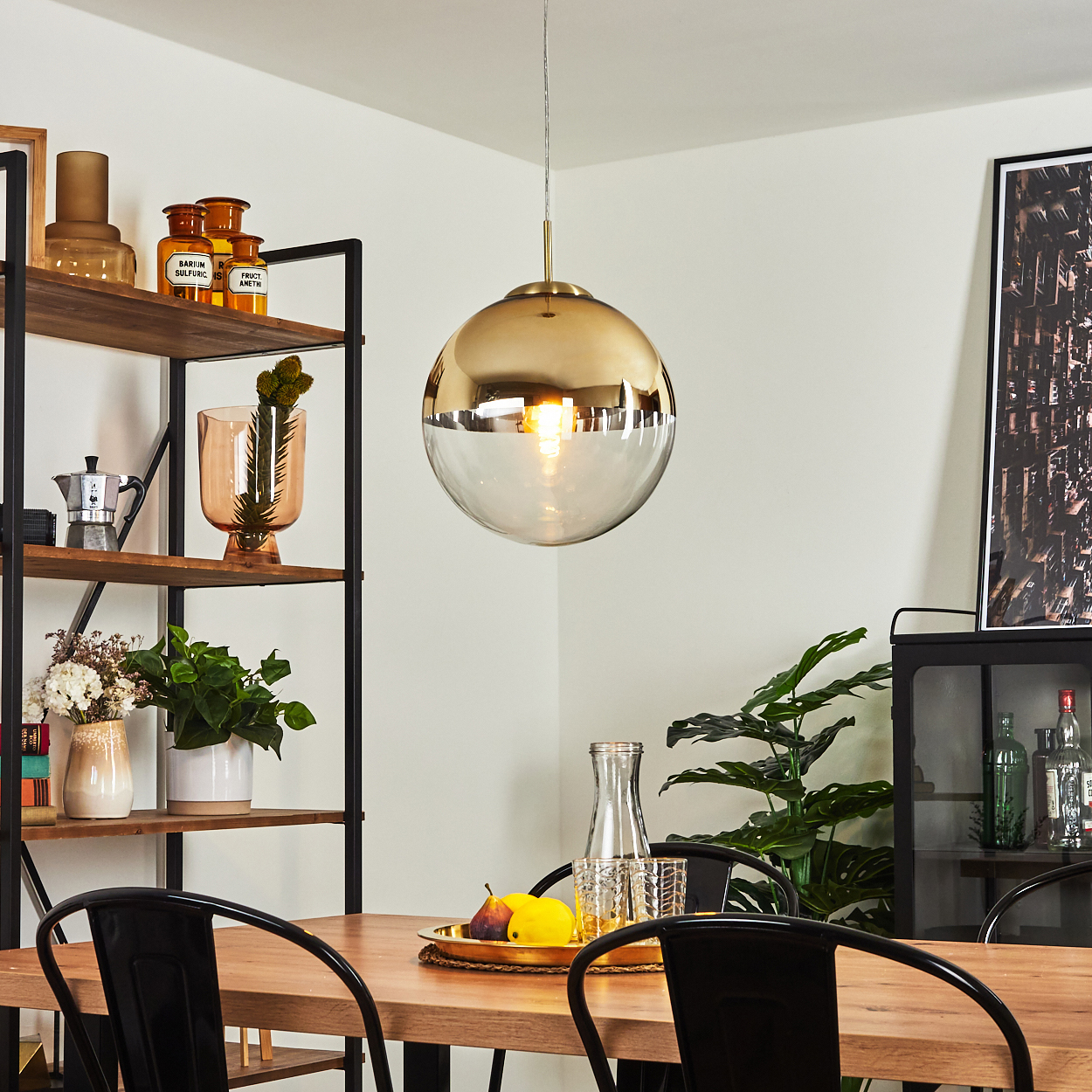 Esszimmer-Szene. Eine goldene und gläserne Kugellampe hängt über einem Holztisch mit Obst und schwarzen Stühlen. Regale mit Dekoration sind sichtbar.