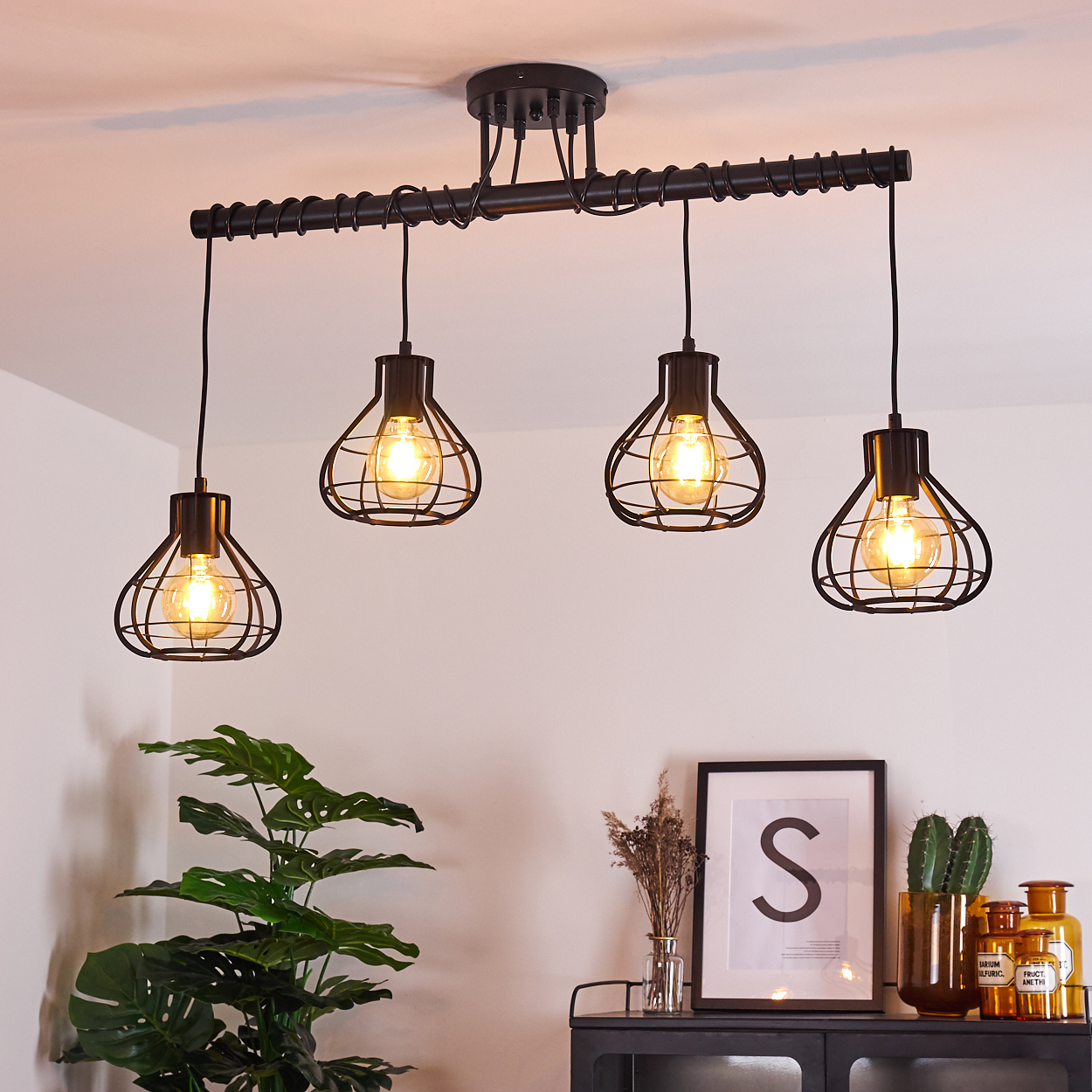 Vier schwarze Pendelleuchten mit käfigartigen Glühbirnen hängen über der Einrichtung. Eine Pflanze, gerahmte Kunst mit einem „S“ und Flaschen stehen auf einem Schrank.