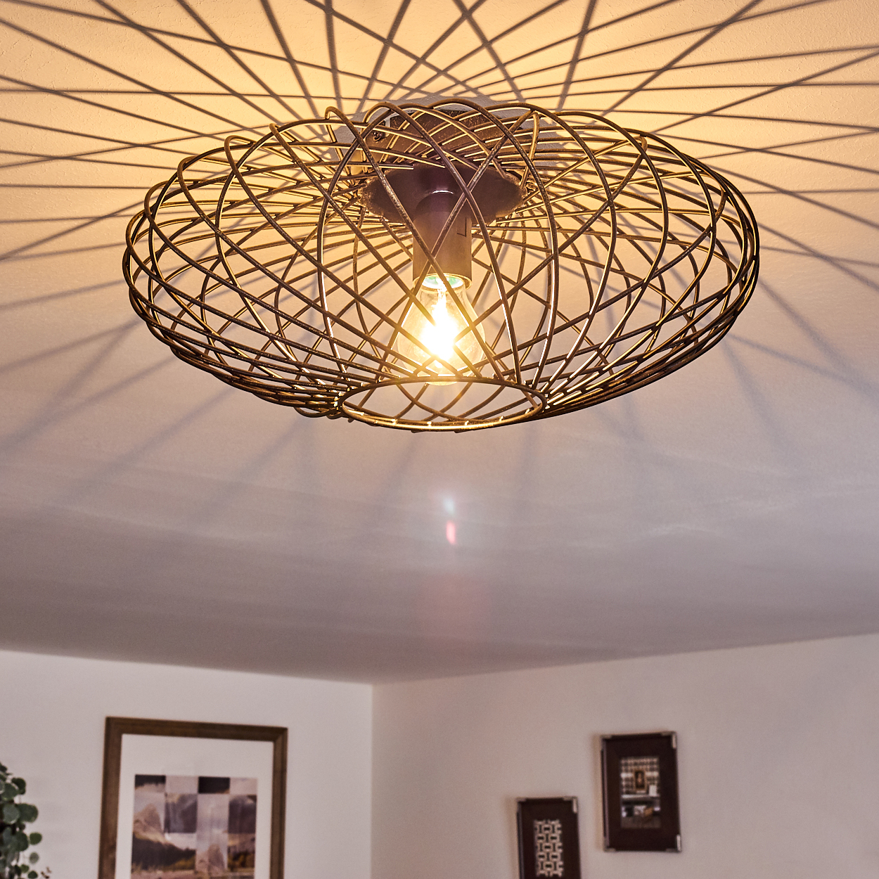 Eine ovale Metallkäfig-Leuchte an einer weißen Decke. Das Licht ist an. Schatten des Käfigs werden auf die Decke geworfen.
