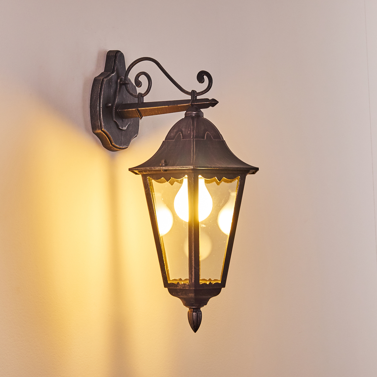 Eine Außenwandleuchte mit dunklem Metallrahmen und Glasscheiben, beleuchtet von hellen Lichtern an einer cremefarbenen Wand.