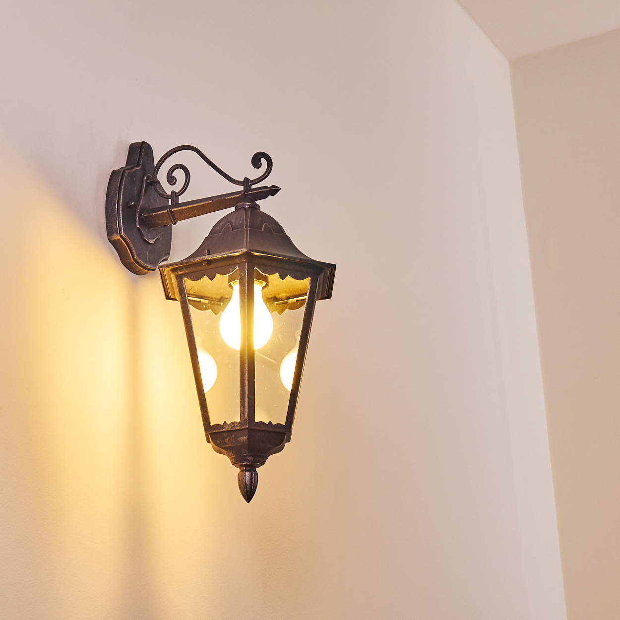 Eine Wandlaterne mit einer leuchtenden Glühbirne. Dunkles Metall vor einer hellen Wand, die ein warmes Licht wirft.