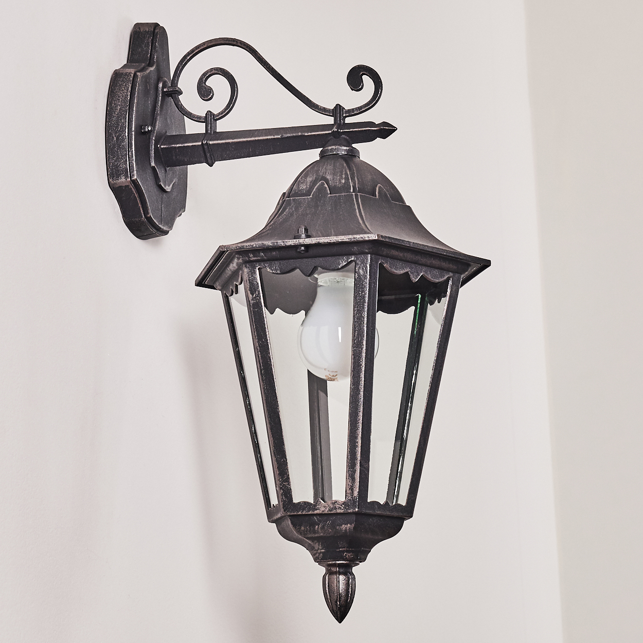 Eine schwarze, verzierte Außenlampe ist an einer weißen Wand montiert. Sie hat ein Glasgehäuse mit einer Glühbirne.