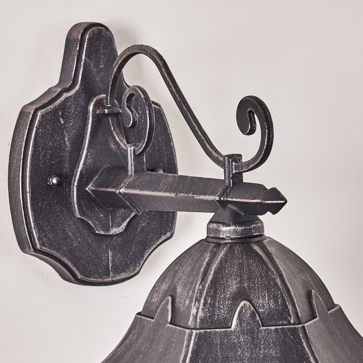Eine an der Wand befestigte, verzierte Metalllampe. Sie hat eine dekorative Halterung, einen gebogenen Arm und einen glockenförmigen Schirm.