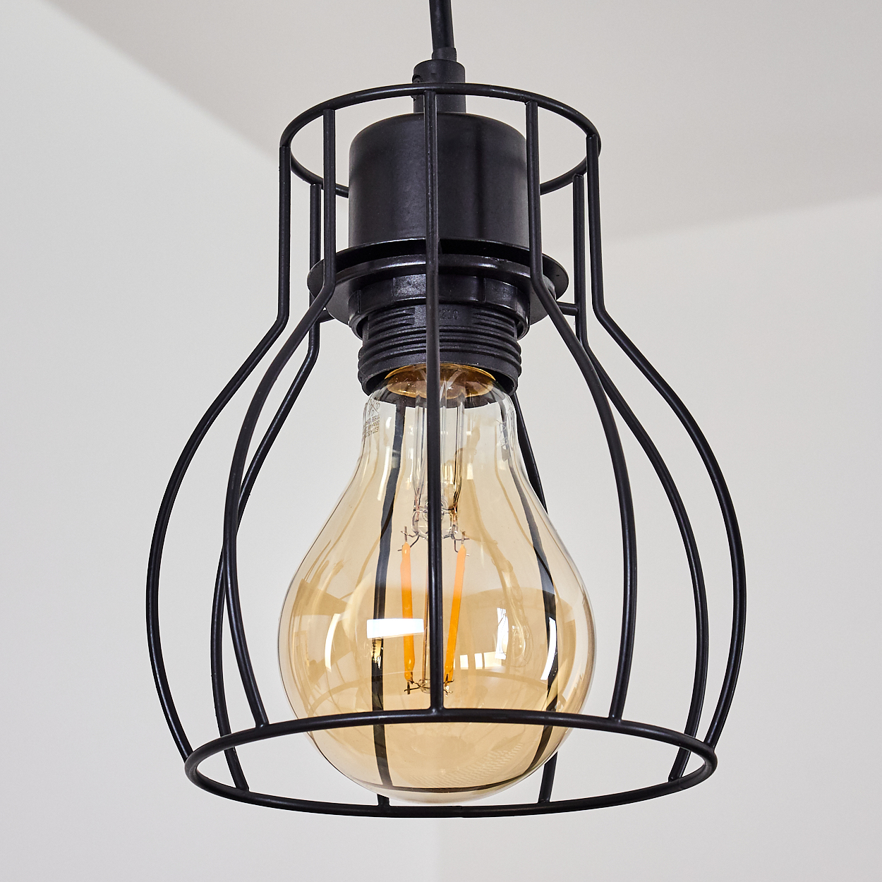 Eine schwarze Metallkäfig-Leuchte mit einer großen, bernsteinfarbenen Edison-Glühbirne. Der Hintergrund ist eine schlichte weiße Decke.