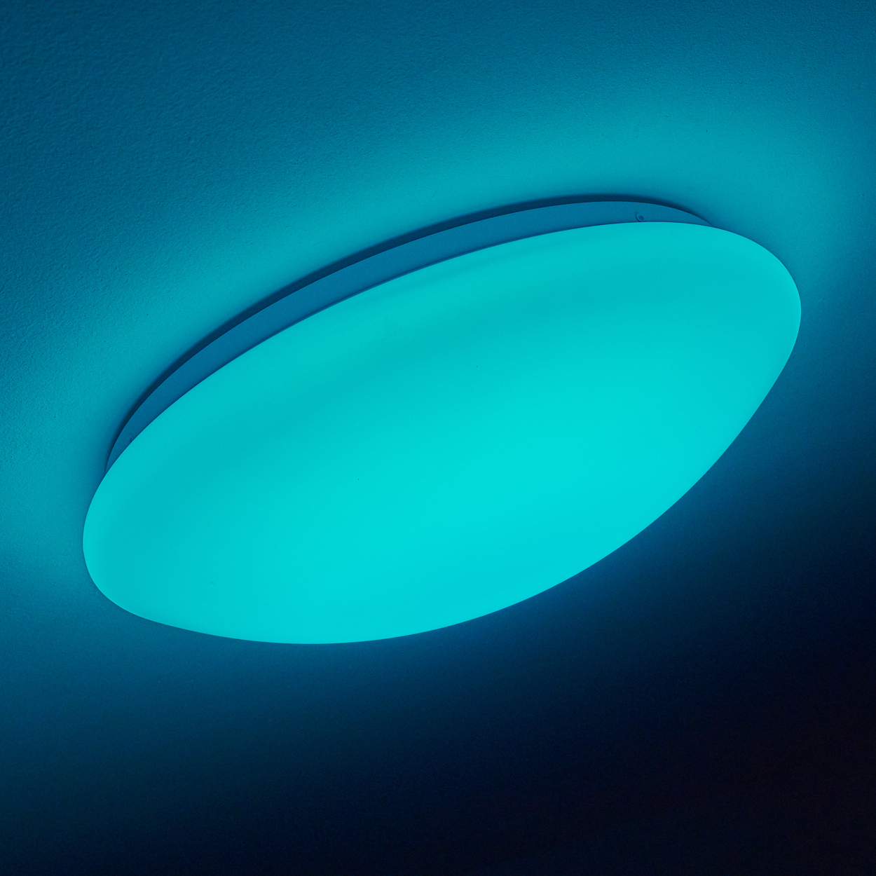 Eine ovale, türkis gefärbte Deckenleuchte vor einer dunkelblauen Decke. Das Licht ist eingeschaltet.