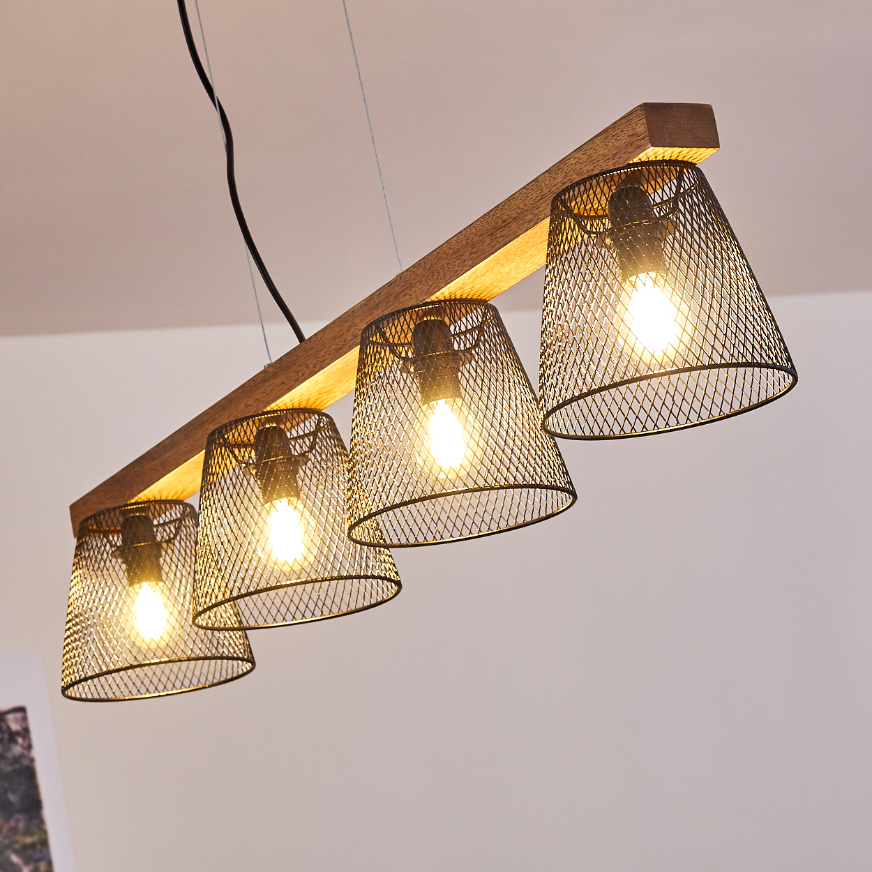 Ein Holzbalken mit vier Lampenschirmen aus Metallgeflecht, jede mit einer brennenden Glühbirne. Der Hintergrund ist eine weiße Decke.