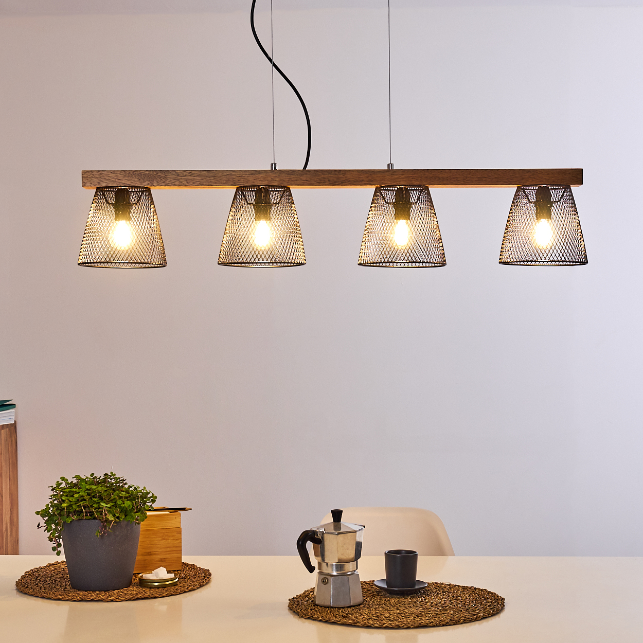 Ein Holzbalken mit vier Mesh-Lampen hängt über einem Tisch. Auf dem Tisch stehen eine Pflanze, eine Kaffeemaschine und eine Tasse.