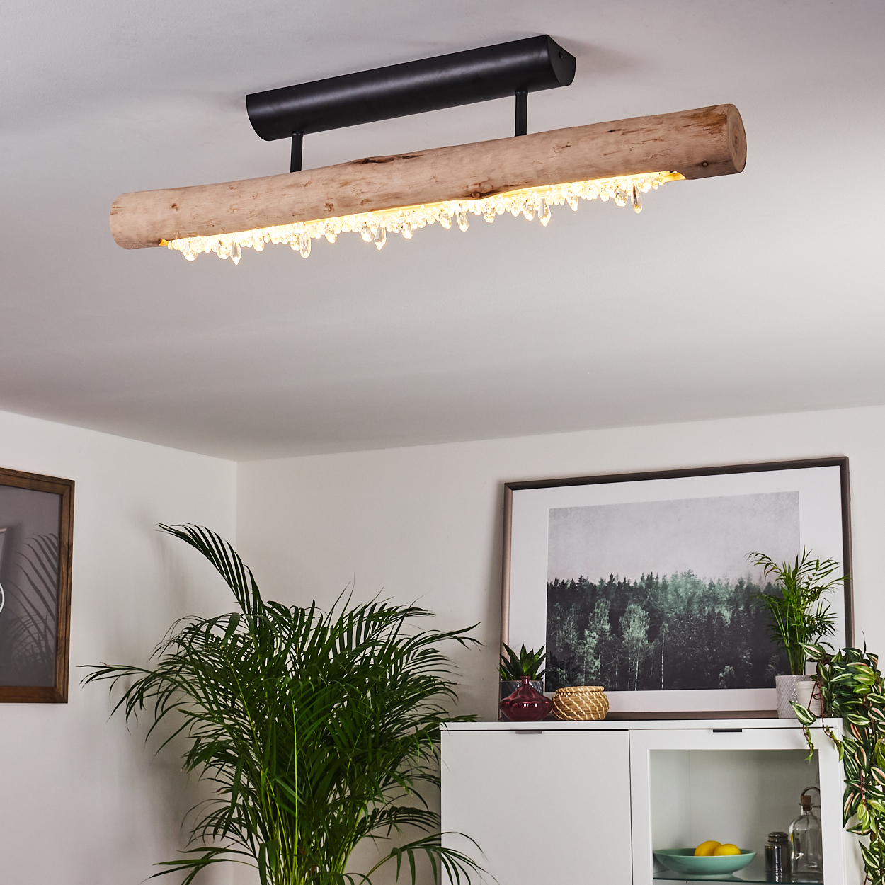 Eine Holz- und Kristall-Kronleuchter hängen von der Decke. Darunter ein weißer Schrank mit einem Waldbild und grünen Pflanzen.