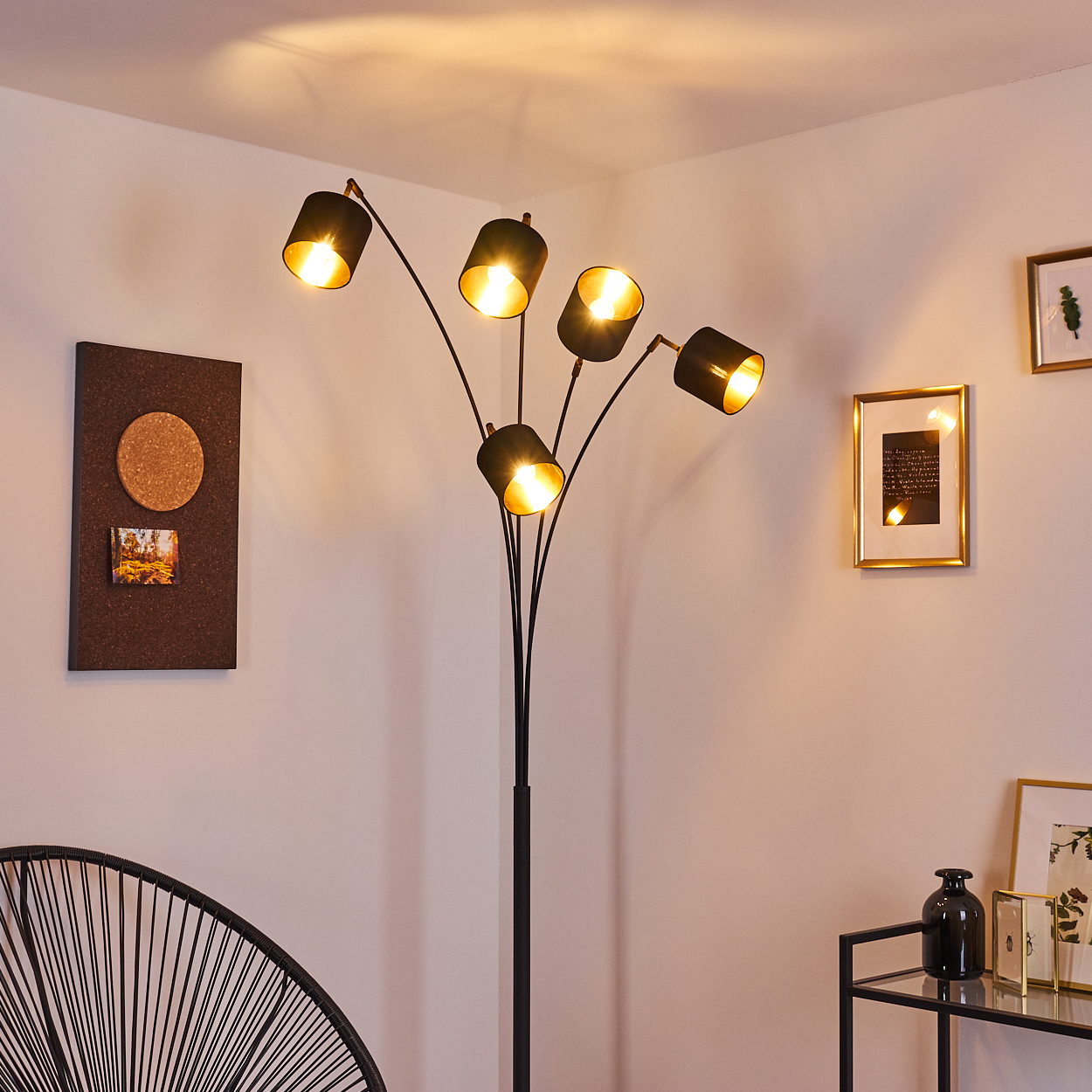 Eine schwarze Stehlampe mit fünf Lichtern beleuchtet einen Raum. Eine Pinnwand und gerahmte Bilder schmücken die weißen Wände.