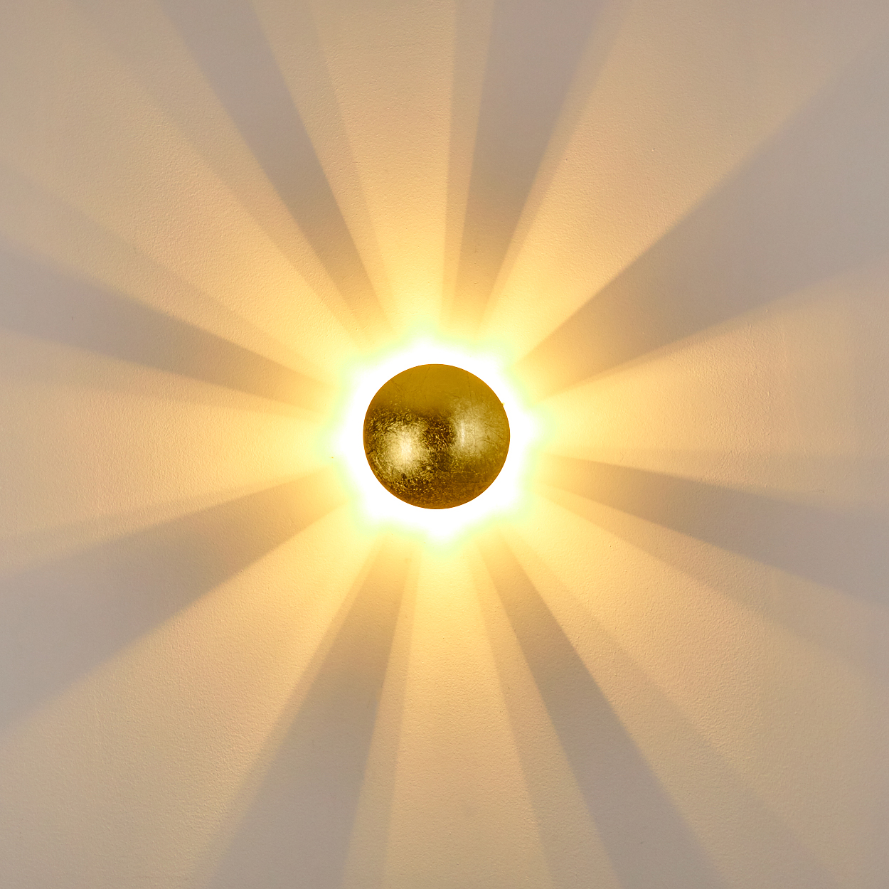 Eine goldene, kugelförmige Leuchte strahlt in einem Sonnenstrahlenmuster gegen eine strukturierte, weiße Wand. Die Lichtstrahlen sind sichtbar.