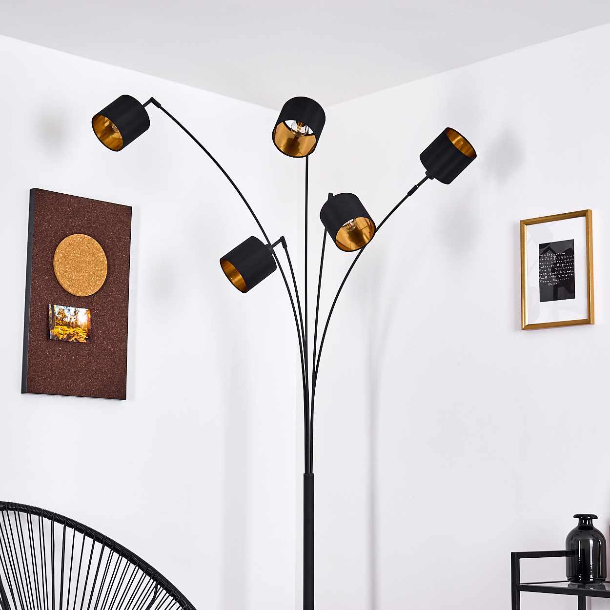 Eine schwarze Stehlampe mit fünf Schirmen, goldenen Innenseiten, vor einer weißen Wand. Eine Pinnwand, ein gerahmtes Bild und ein schwarzer Stuhl sind zu sehen.