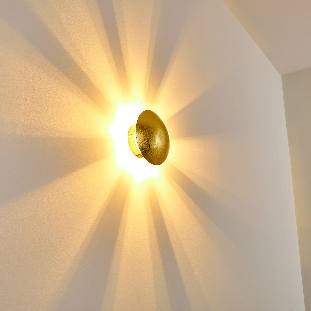 Eine goldene Wandleuchte an einer weißen Wand. Licht strahlt aus und erzeugt Schatten. Die Lampe ist eiförmig.