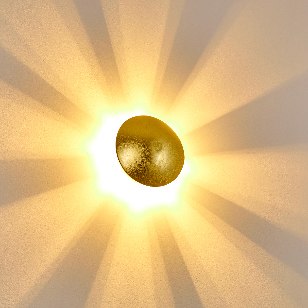 Eine goldene, ovale Wandleuchte strahlt helles Licht aus und wirft radiale Schatten auf eine strukturierte, cremefarbene Wand.