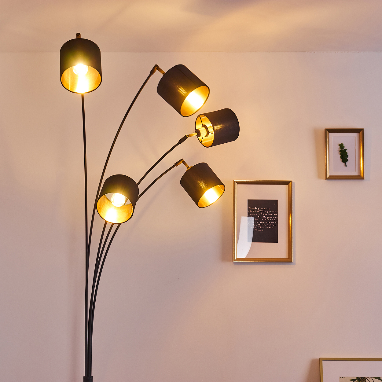 Eine schwarze Stehlampe mit fünf Lampenschirmen beleuchtet eine beige Wand. Zwei gerahmte Bilder hängen an der Wand.