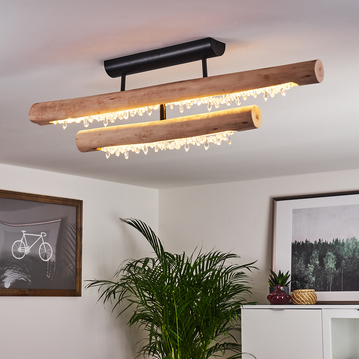 Ein Holz-Kronleuchter mit Kristallen hängt von der Decke. Darunter befinden sich eine Pflanze und Bilder. Der Raum hat eine weiße Wand.